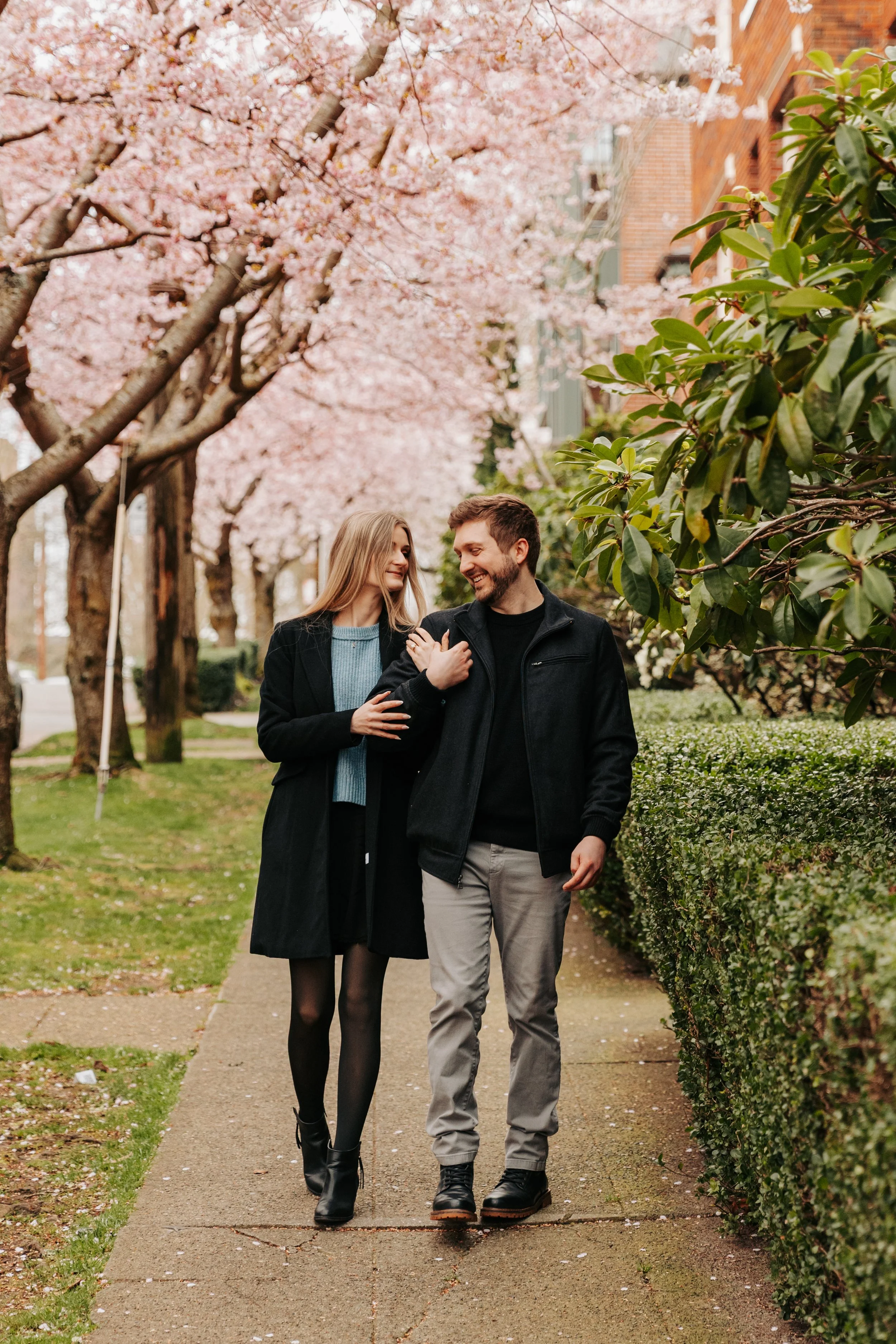 A young woman and man walking together on a sidewalk lined with blooming pink cherry blossom trees and green bushes, smiling and enjoying each other's company.