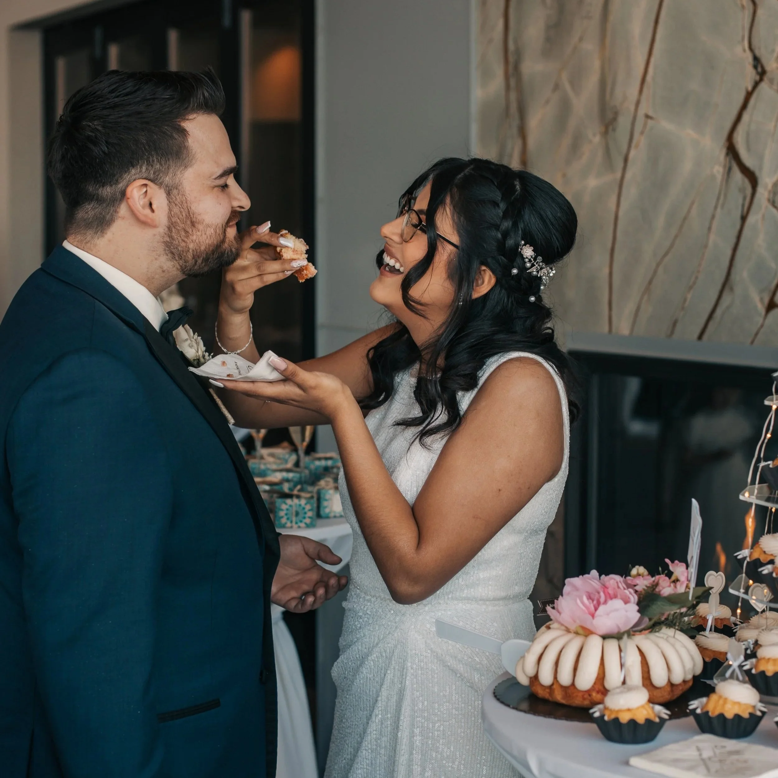 A woman in a wedding dress feeding a slice of cake to a man in a tuxedo at a wedding reception, with a dessert table with cupcakes and flowers in the background.