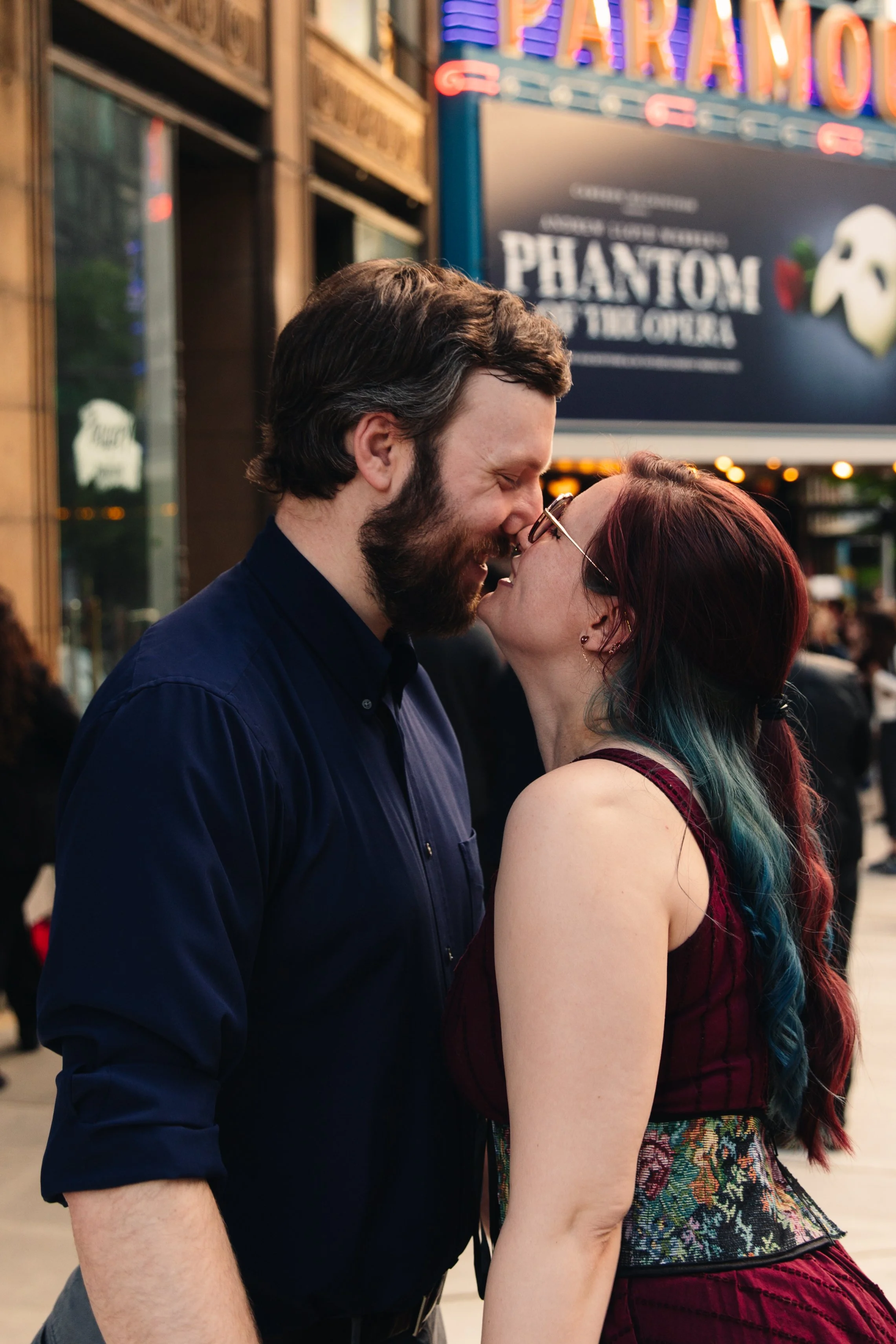A man and woman are standing close, about to kiss, in front of a theater marquee for 'Phantom of the Opera'.