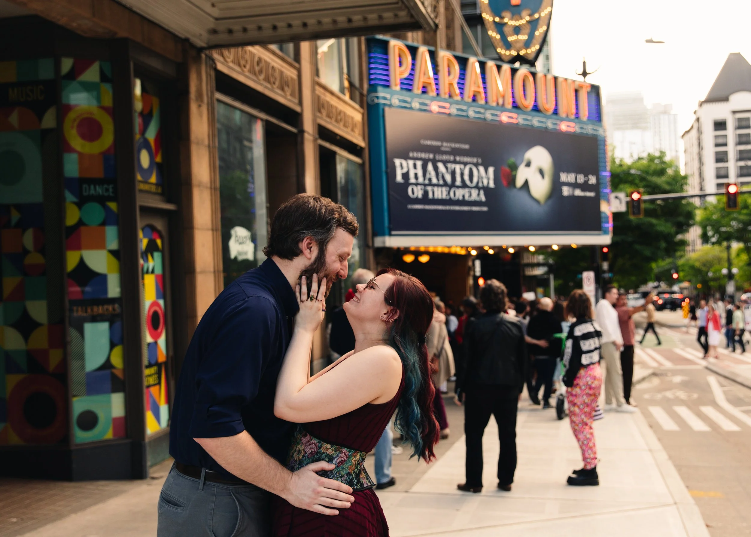A couple with their foreheads and noses touching, sharing an intimate moment on a busy city street with a theater marquee for 'Phantom of the Opera' in the background.