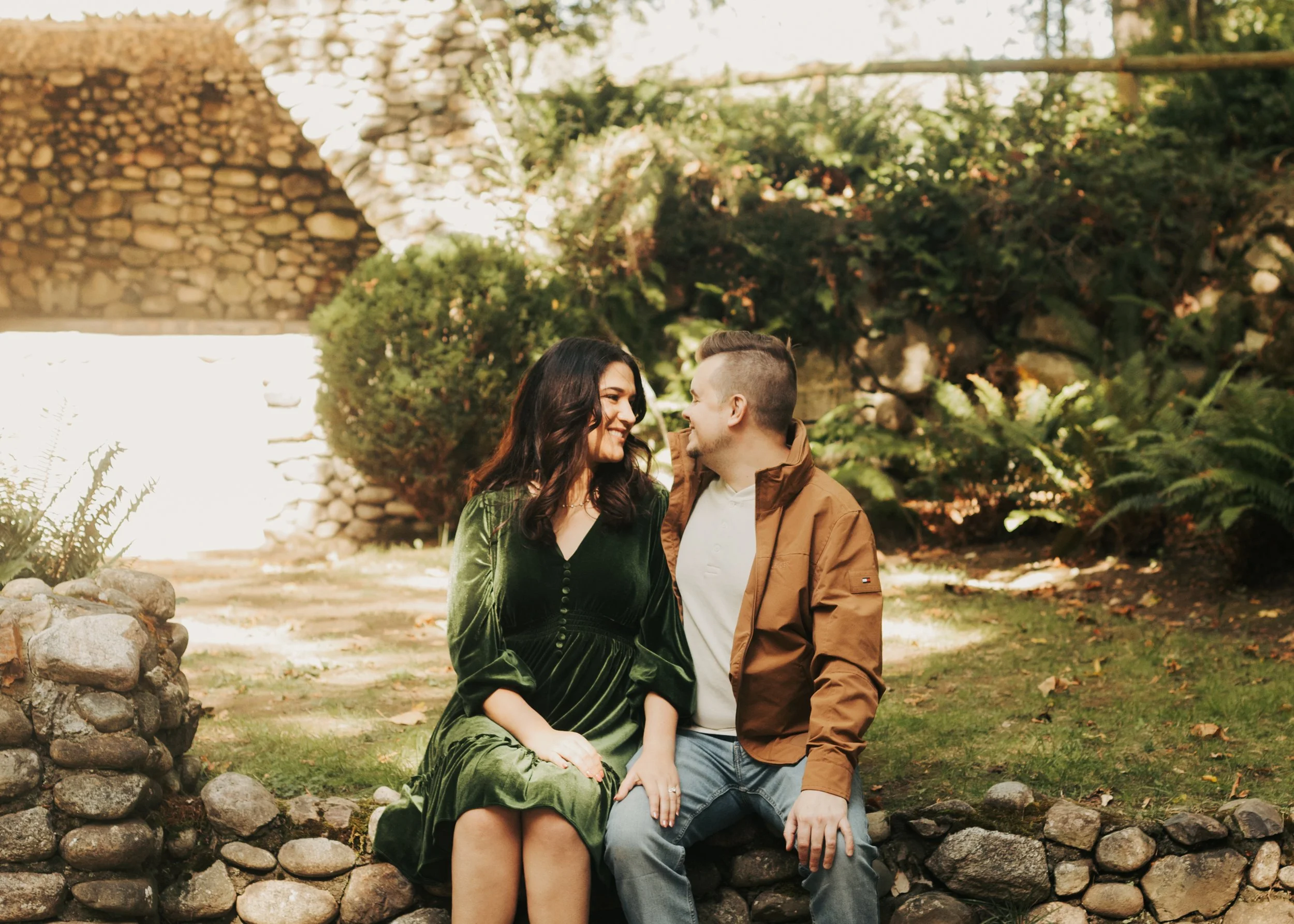 A couple sitting on a stone wall in a garden, smiling and looking at each other with greenery and a stone wall in the background. St. Edwards State Park Washington engagement