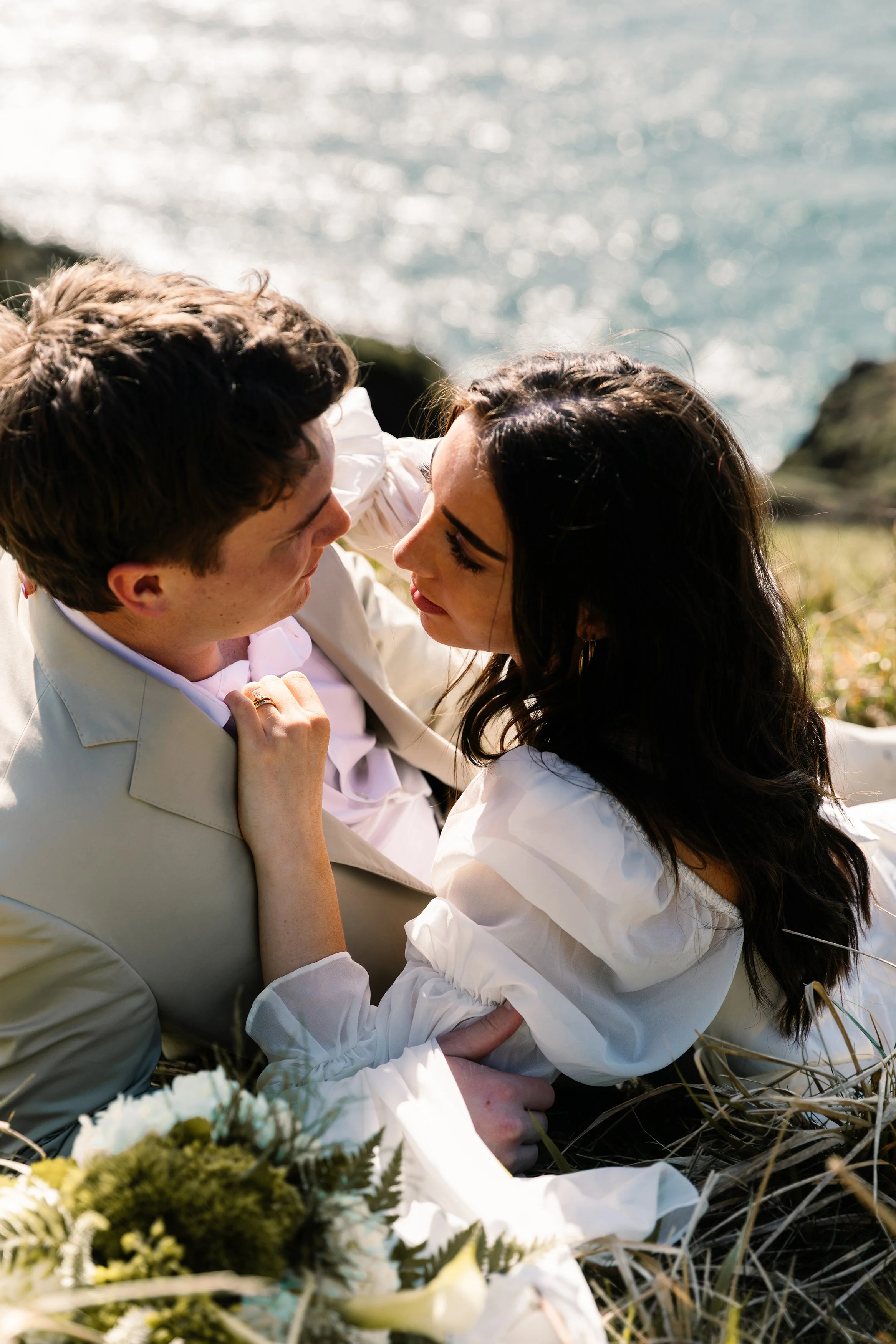 A couple lying on grass near the ocean, gazing into each other's eyes, with flowers nearby, on a sunny day. Elk Flats Oregon elopement