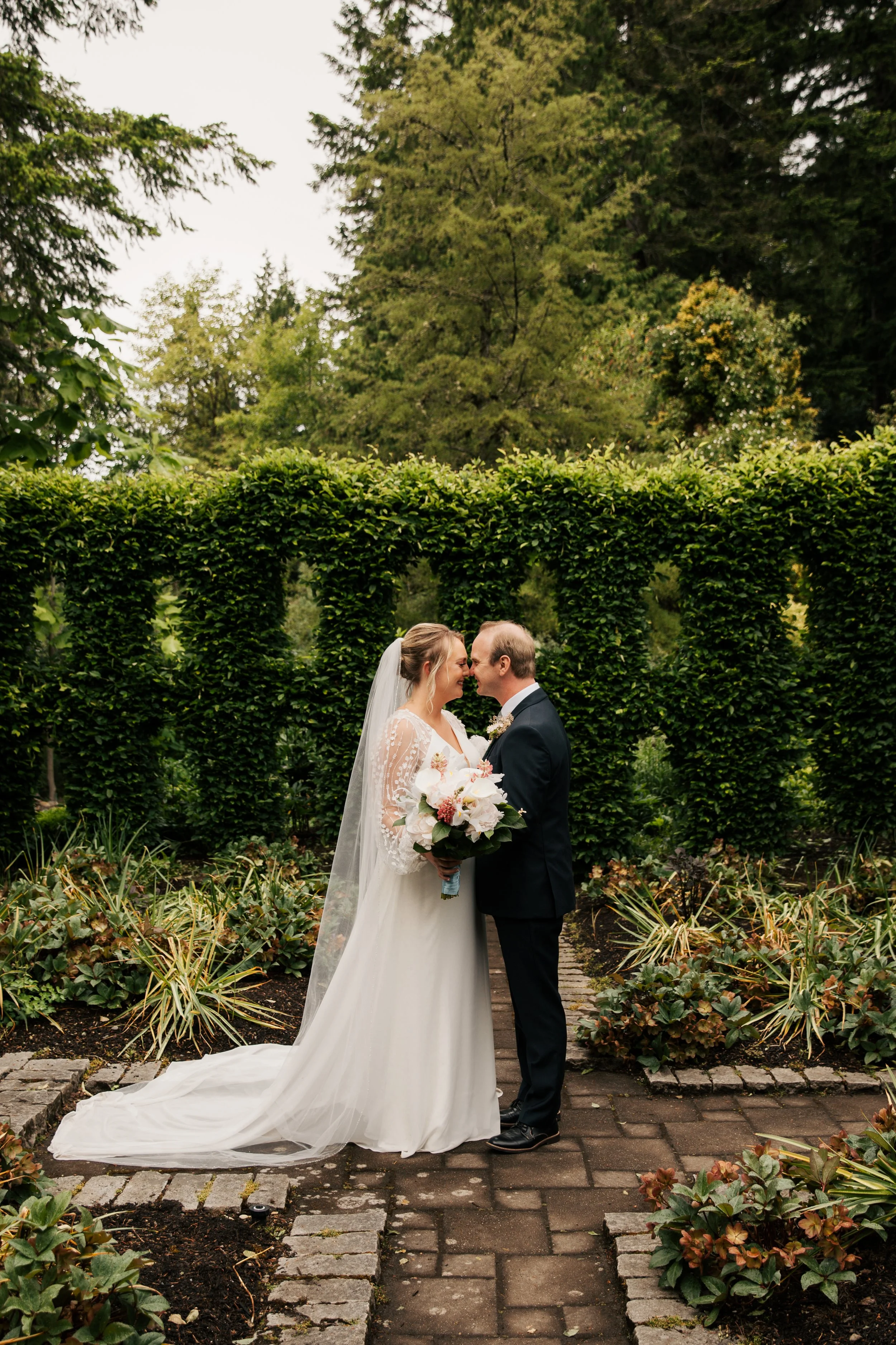 Heronswood Garden wedding Kingston Washington. The bride is in a white gown with lace sleeves, holding a bouquet, and wearing a veil. The groom is in a dark suit. Green plants and trees surround them.