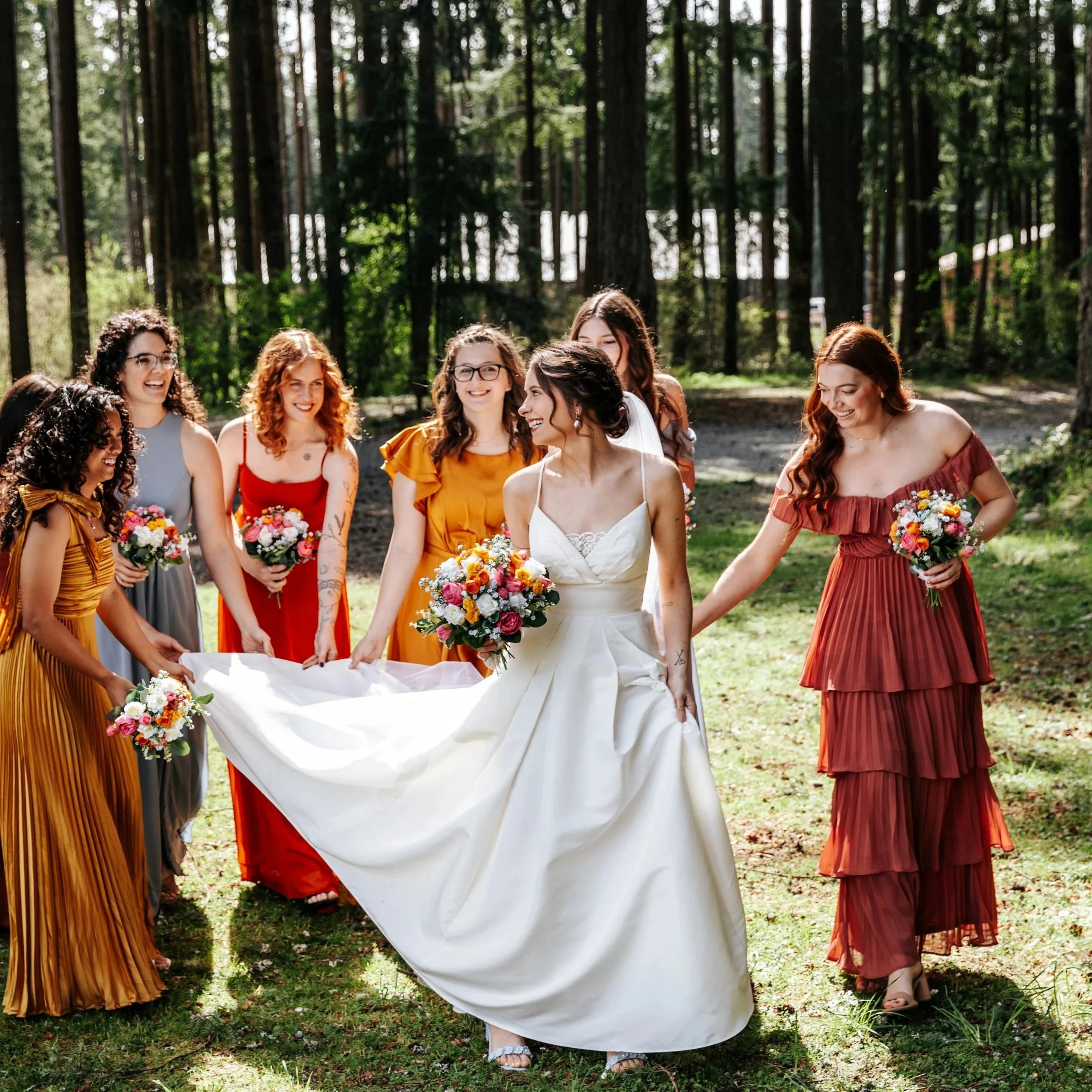 A bride in a white wedding dress holding a bouquet, walking in a forested outdoor setting with seven bridesmaids dressed in colorful dresses, all holding bouquets and smiling. Frontier Park Graham Washington