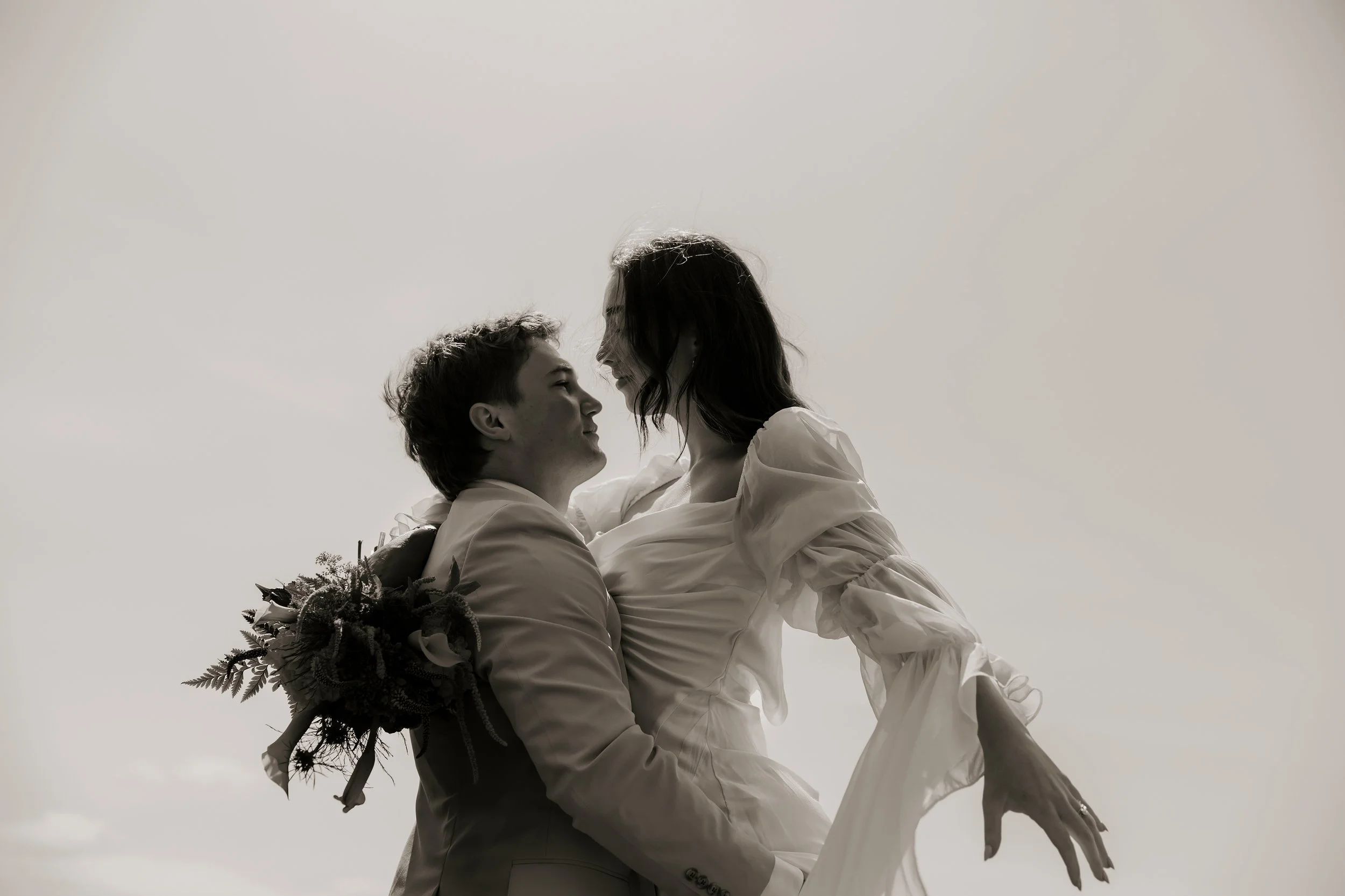 A black and white photo of a man lifting a woman in a dress, both smiling and looking at each other, with the man holding a bouquet of flowers. Elk Flats Oregon