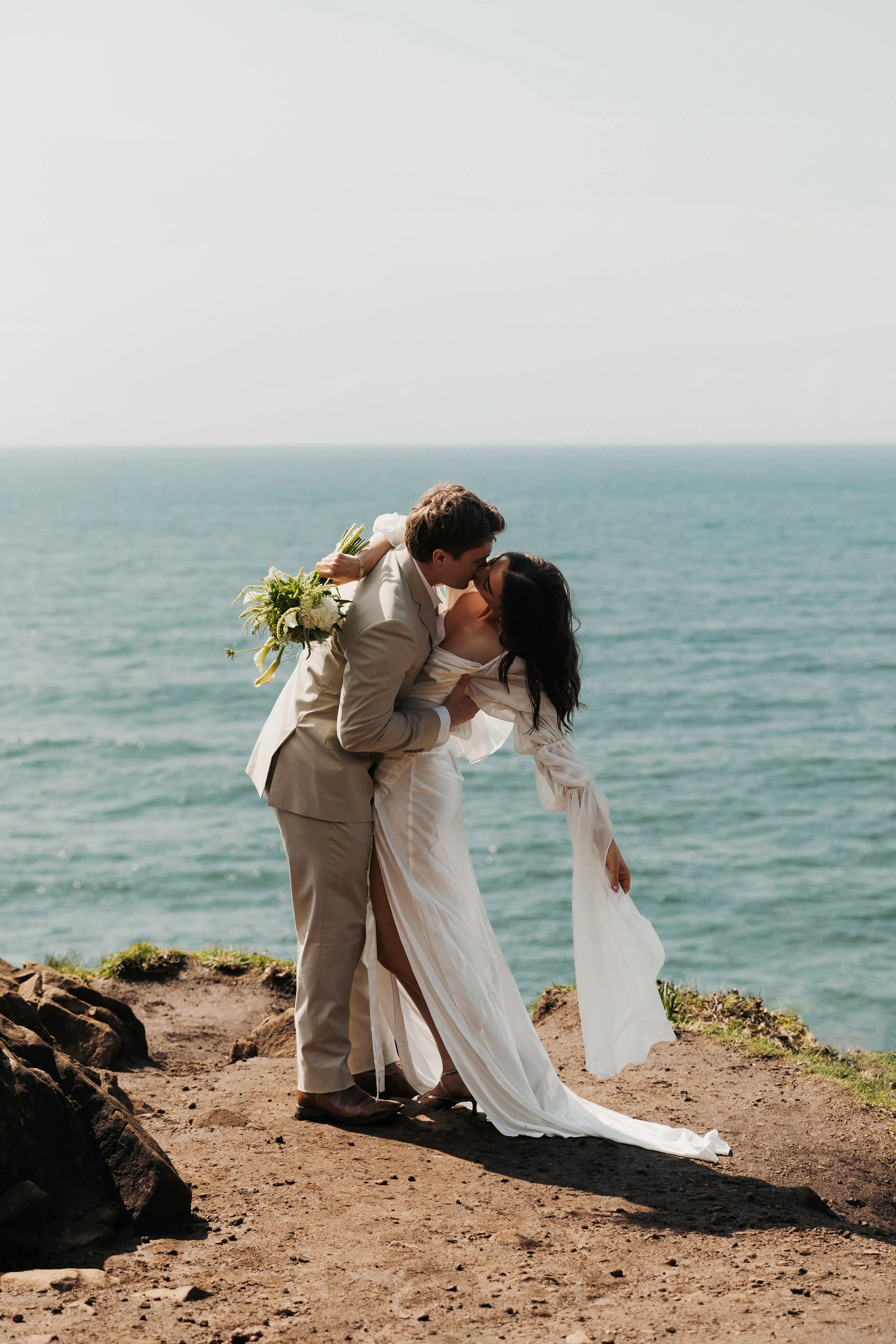A couple in wedding attire sharing a kiss on a cliffside overlooking the ocean. Elk Flats Oregon elopement