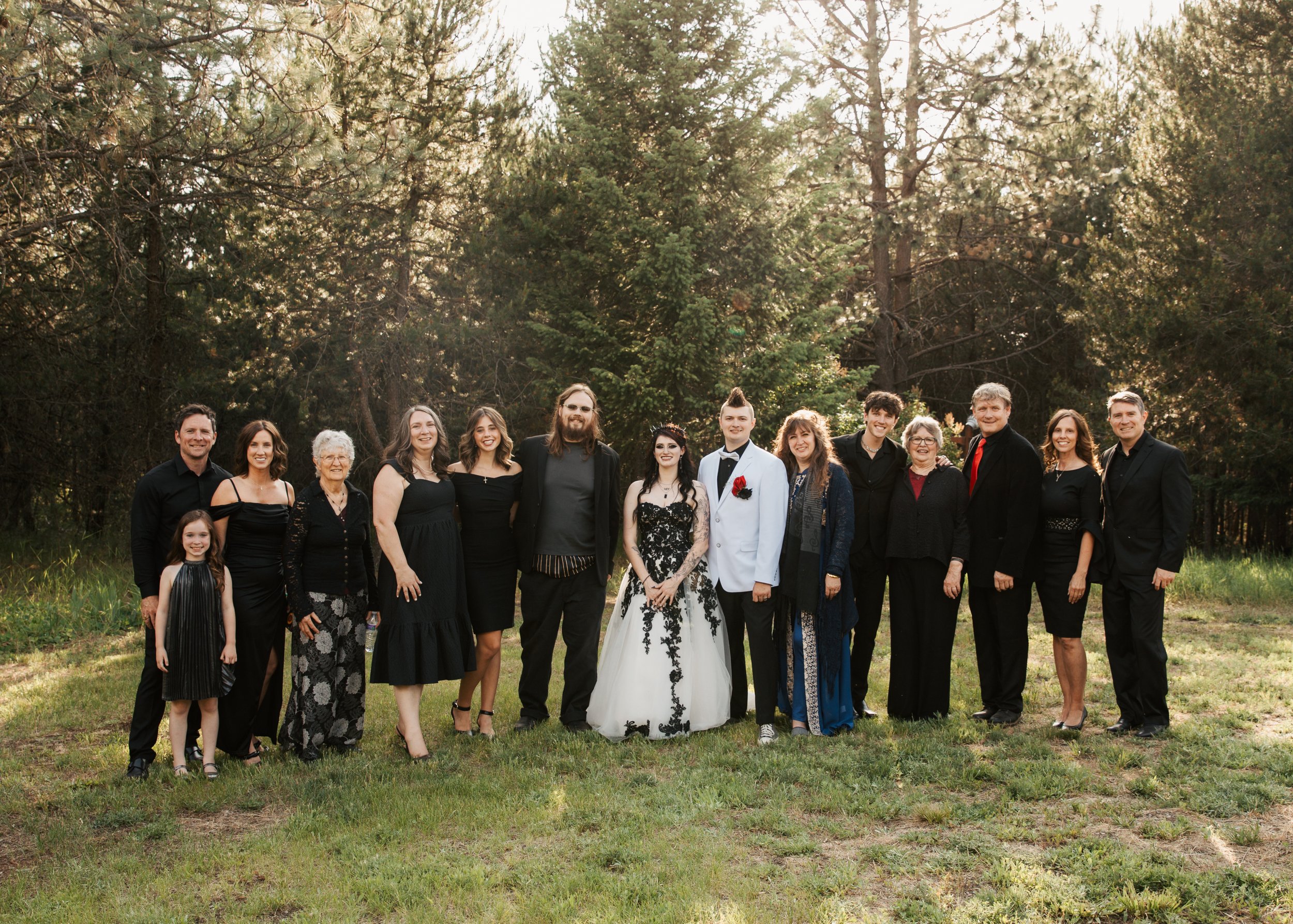 A large family group standing outdoors on grass in front of trees, all dressed in formal attire for a wedding celebration. Athol Idaho wedding. Gothic wedding.