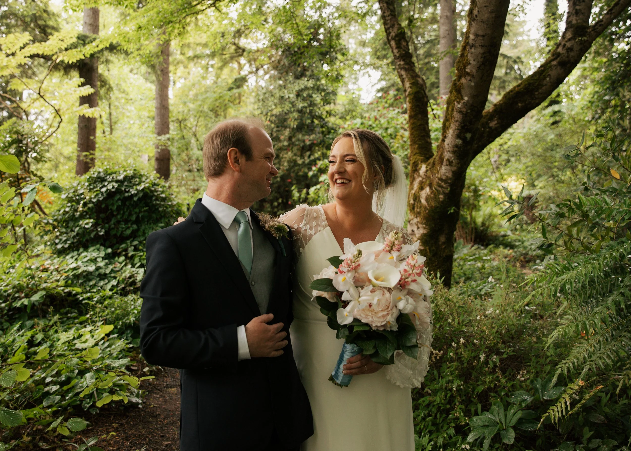 A bride and groom standing together outdoors among green trees and shrubs, smiling at each other, with the bride holding a bouquet of white and pale pink flowers. Heronswood Garden wedding Kingston Washington