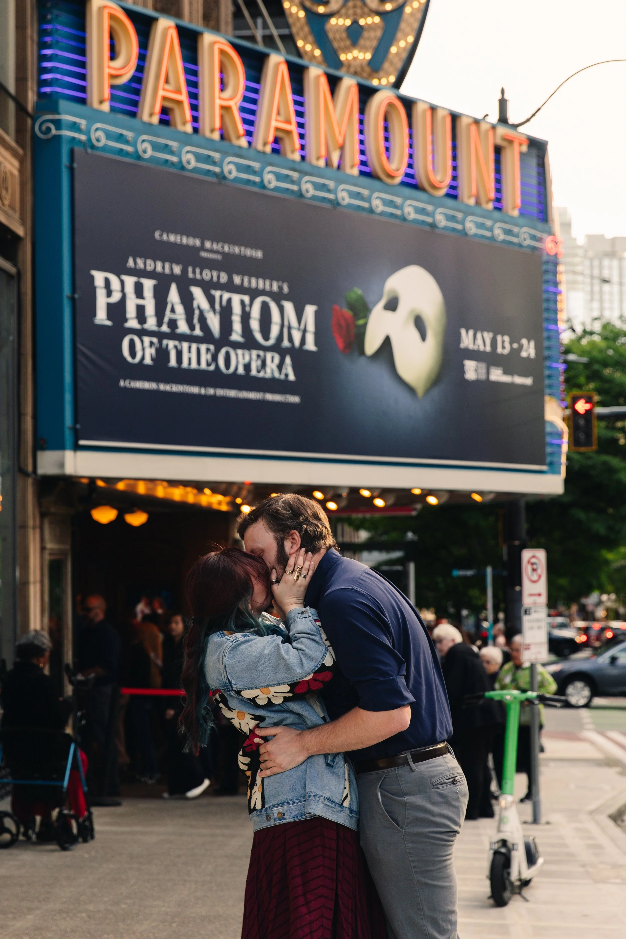 A man and woman embrace in a street scene outside a theater with a marquee for Phantom of the Opera. The man is wearing a dark shirt and gray pants, the woman a patterned jacket and long skirt. They are kissing, surrounded by pedestrians and citylife