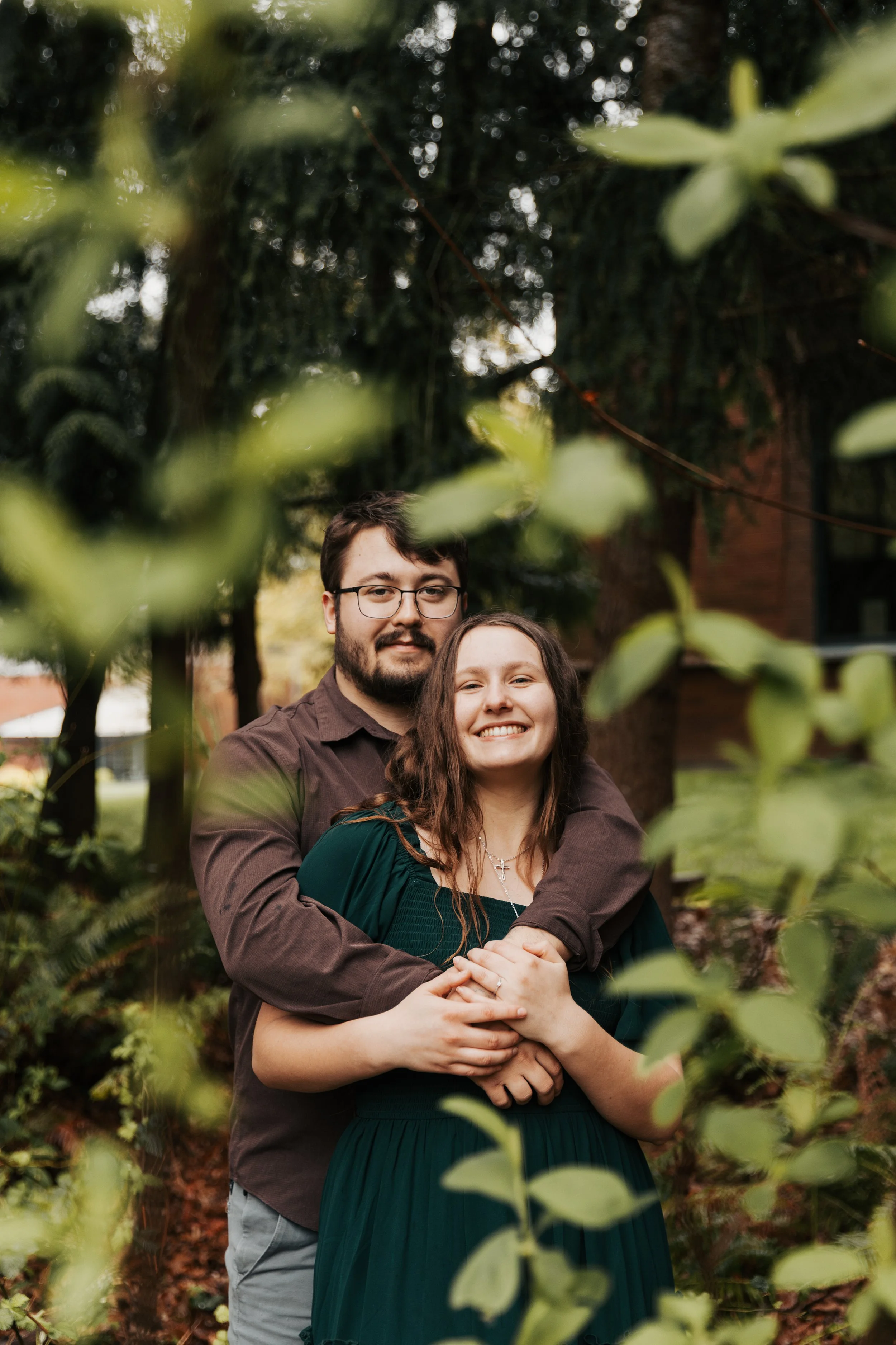 A couple standing outdoors among green leaves, with the man embracing the woman from behind. Both are smiling, and the woman is wearing a dark green dress while the man wears glasses and a brown shirt.