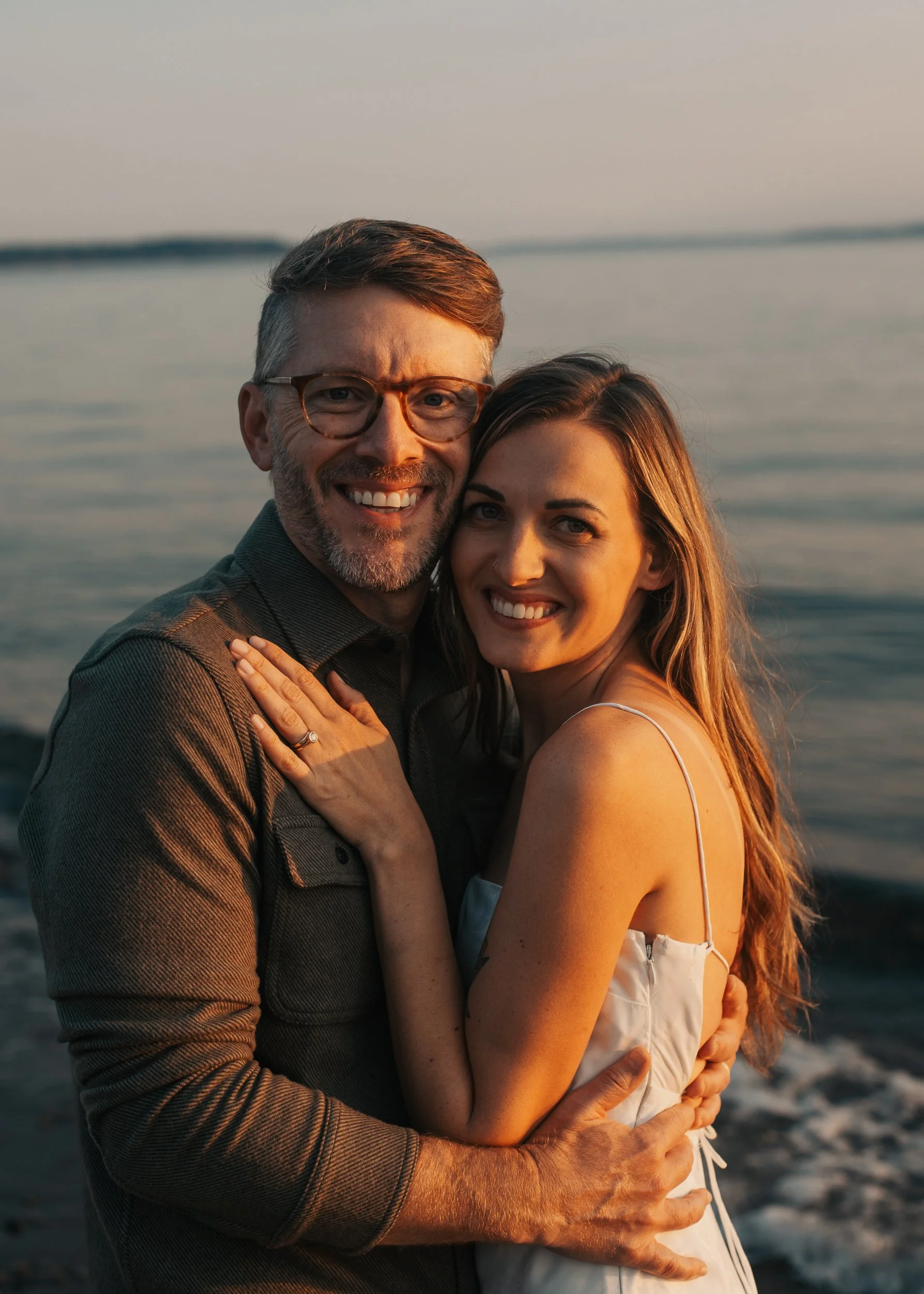 A smiling couple embracing on a beach during sunset, with the ocean in the background. Discovery Park Beach Lighthouse elopement