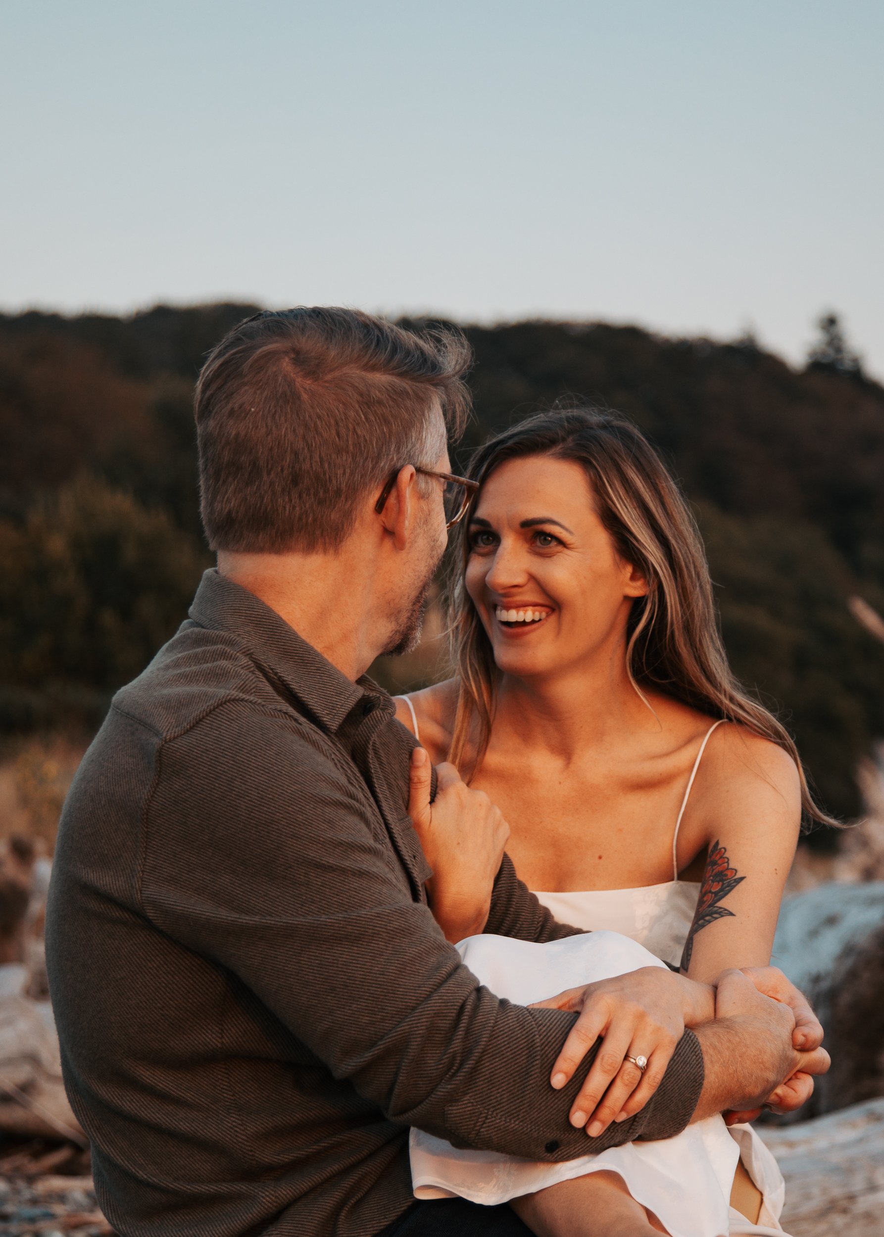 A couple sitting closely together outdoors during sunset, smiling and enjoying each other's company with mountains in the background. Discovery Park Beach Lighthouse elopement