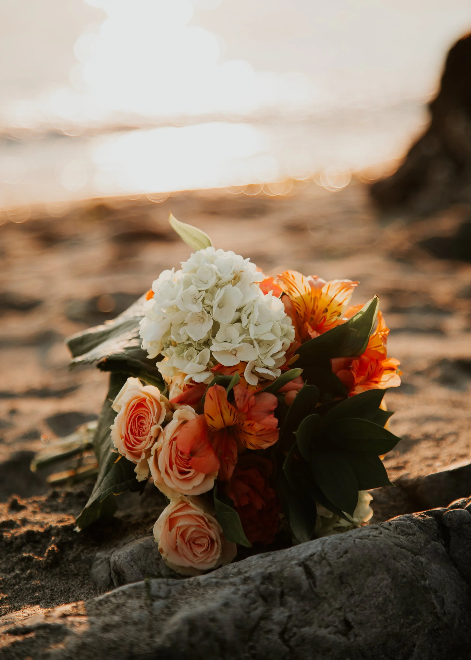 A bouquet of mixed flowers, including white hydrangeas, peach roses, and orange lilies, resting on sand near the ocean with the sunset in the background.