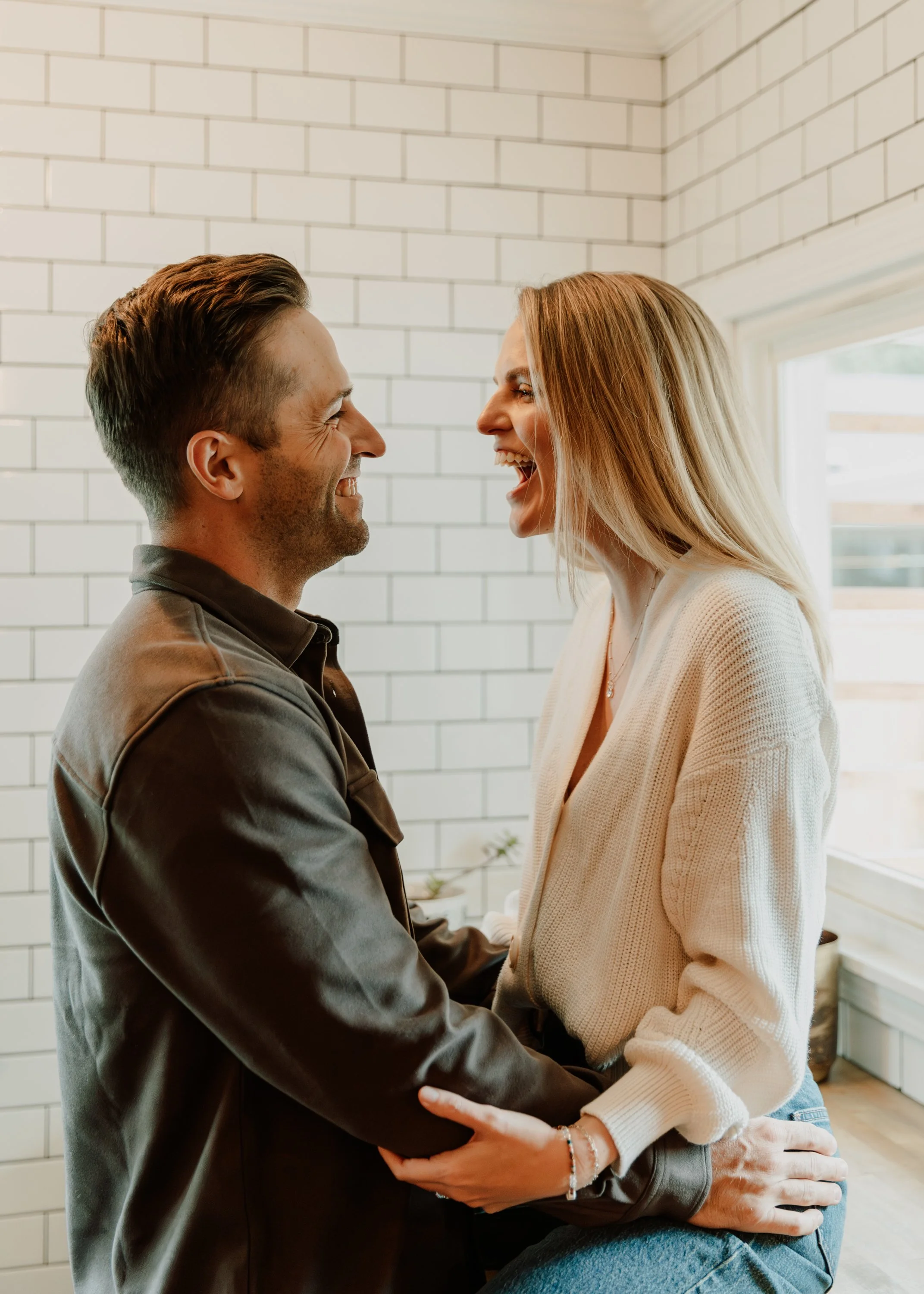 A man and woman are smiling and looking at each other while holding hands in a bright room with white brick walls and a window. Indoor couples photography