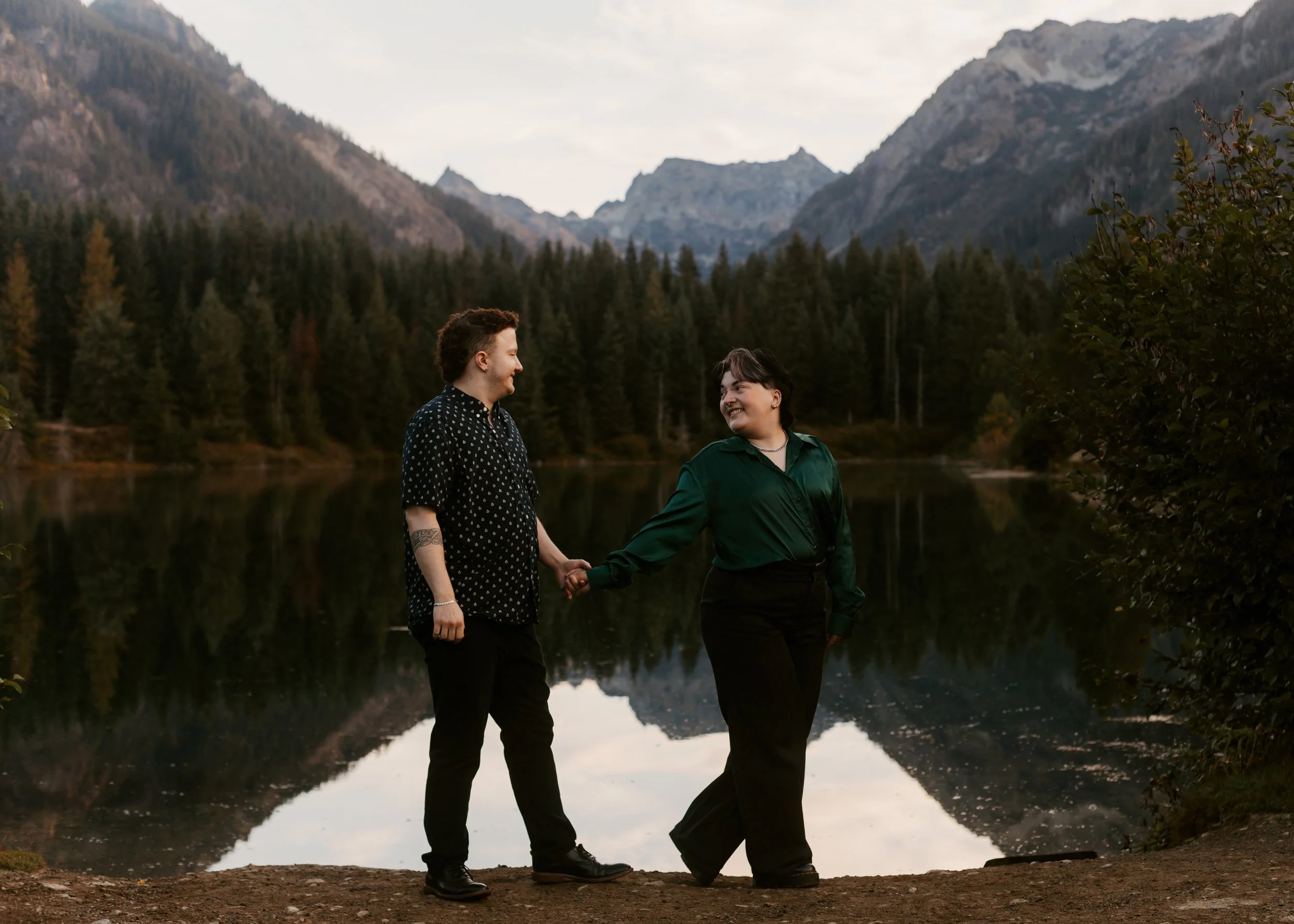 Two people holding hands and smiling at each other near a lake with mountain and forest background. Gold Creek Pond engagement