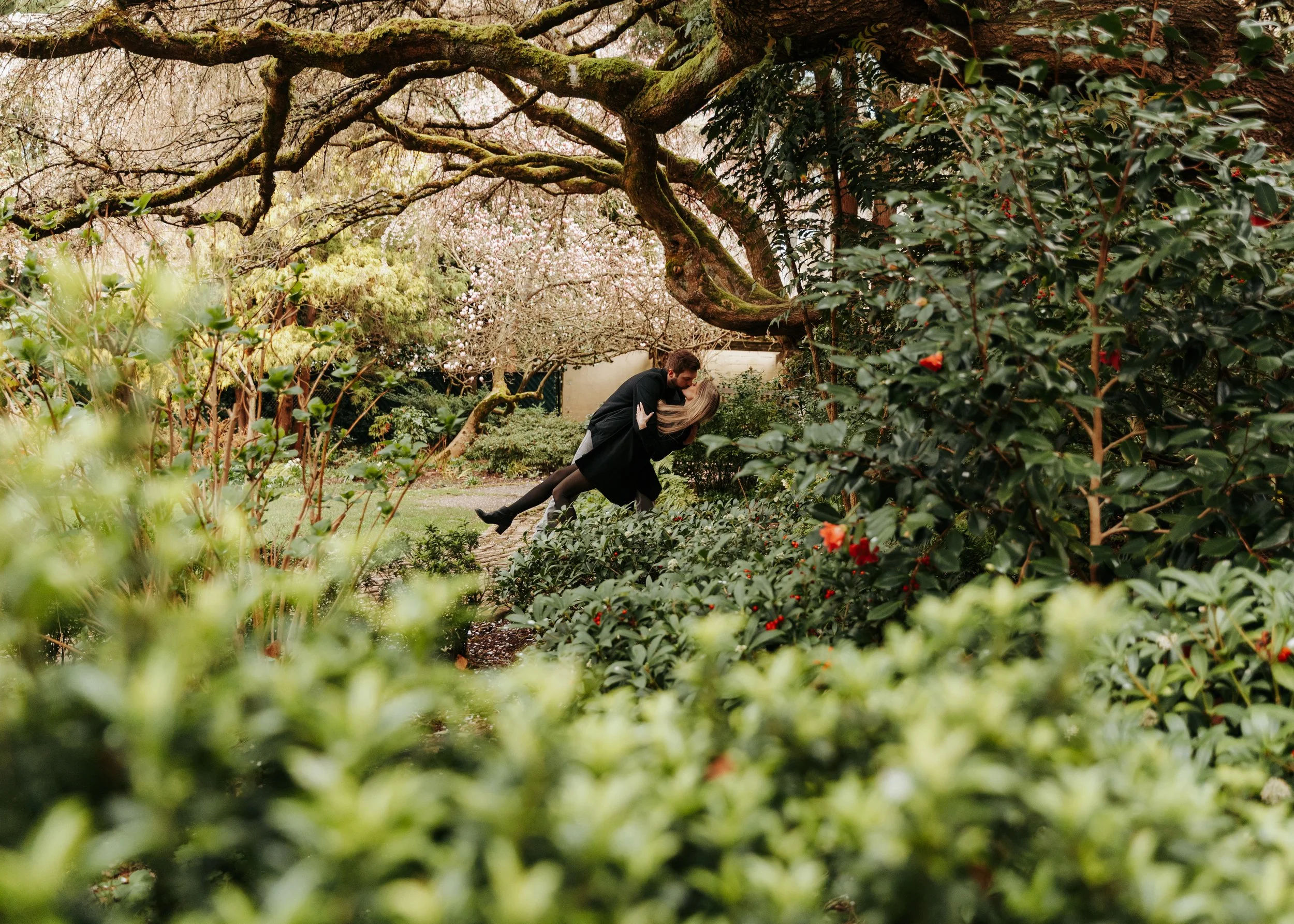 A couple dancing in a lush garden with large trees and flowering bushes.
