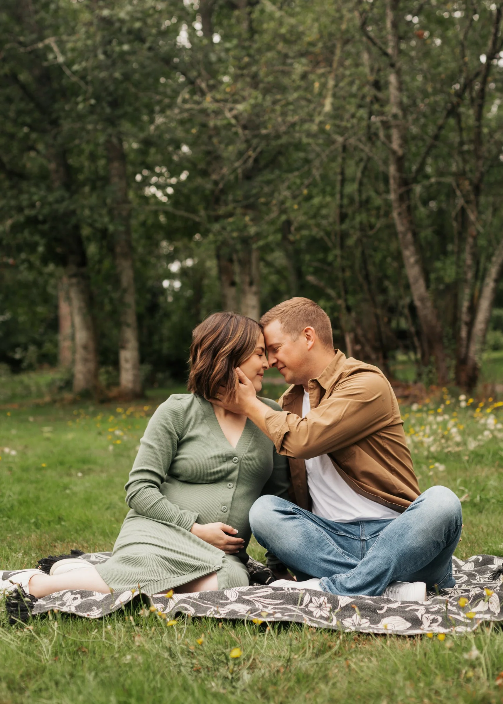 A pregnant woman and a man sitting on a blanket in a park, touching foreheads with eyes closed, surrounded by trees and flowers. Nelson Nature Park Washington