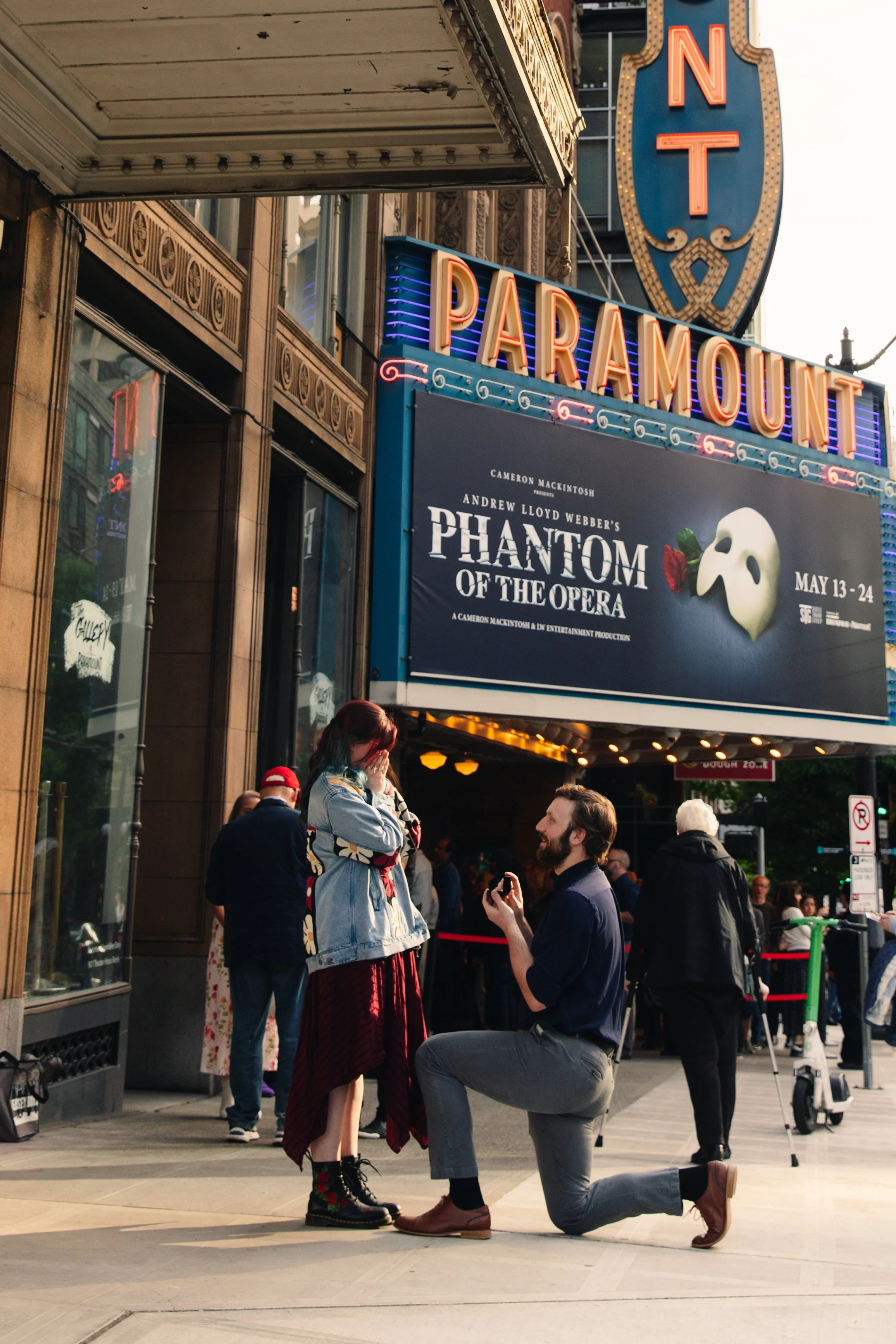A man kneeling on one knee proposing marriage to a woman in front of the historic Paramount Theatre entrance during daytime, with a crowd of onlookers.