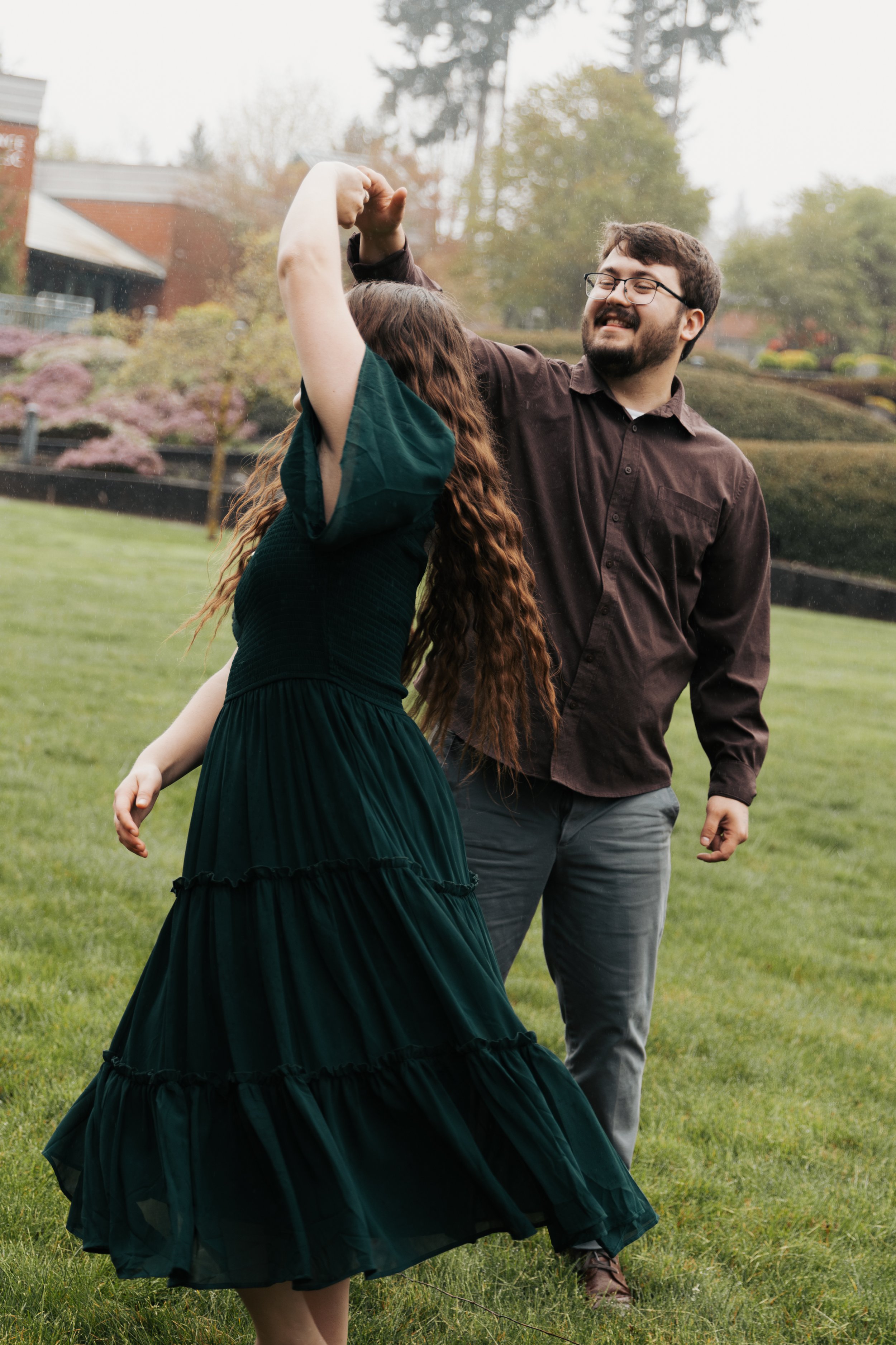 A man and a woman dancing outdoors on a grassy lawn, with trees and houses in the background. The woman is wearing a dark green dress, and the man is wearing a brown shirt and gray pants. They are smiling and holding hands as they dance.