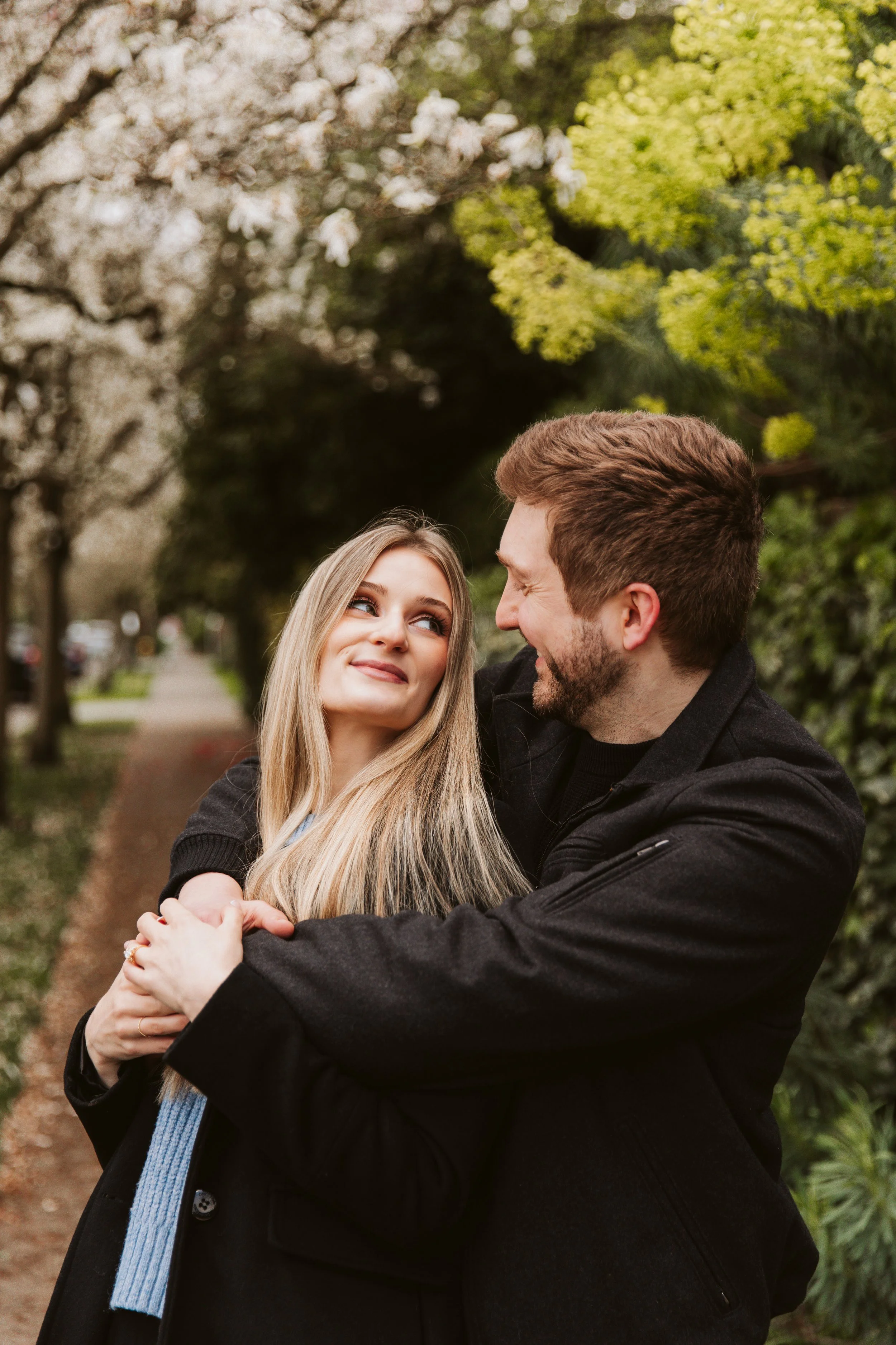 A young couple embracing outdoors among blooming trees, smiling at each other.
