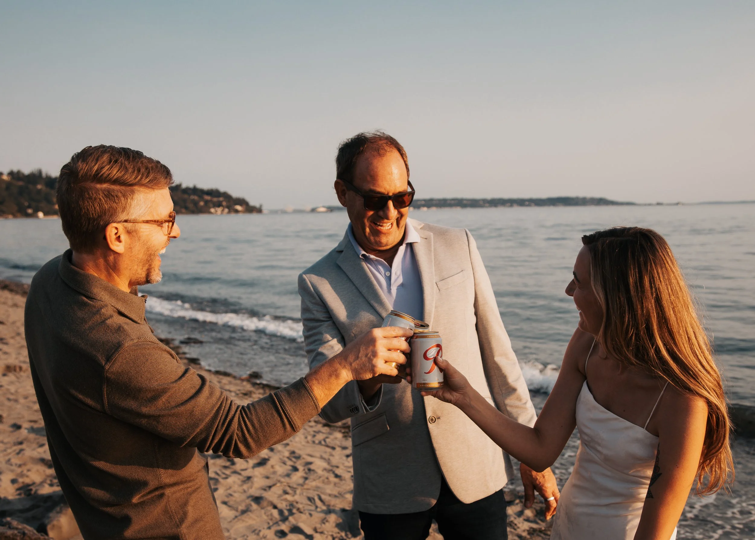 Three people celebrating and sharing drinks on a beach at sunset, with water and distant land in the background. Discovery Park Beach Lighthouse elopement