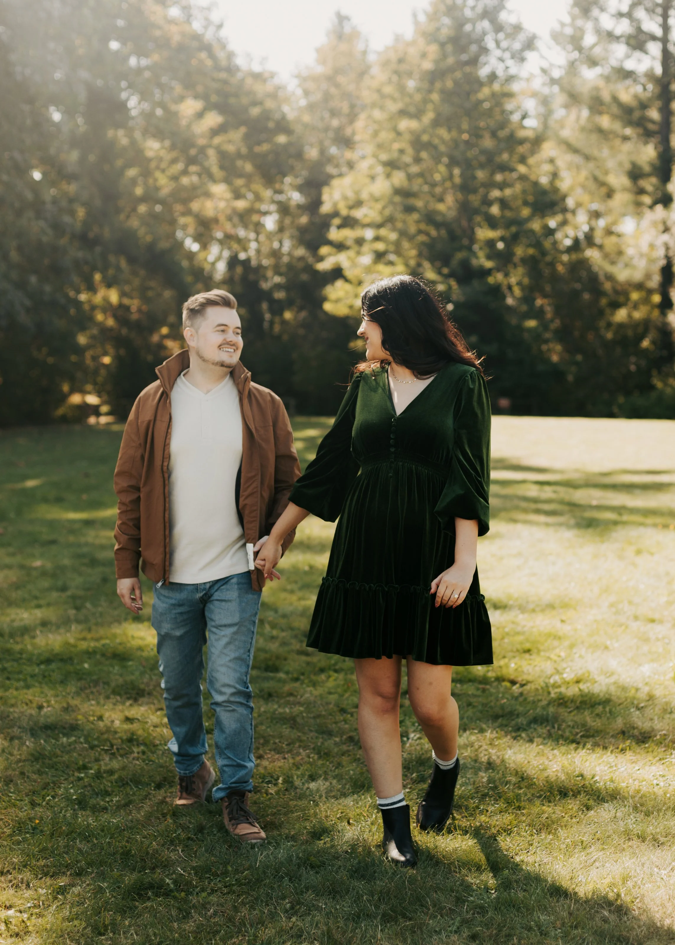 A young man and woman walk hand in hand through a park on a sunny day. The man has short brown hair, a beard, and is wearing a white shirt and brown jacket. The woman has long dark hair, is wearing a dark green dress, black boots, and smiles at the m