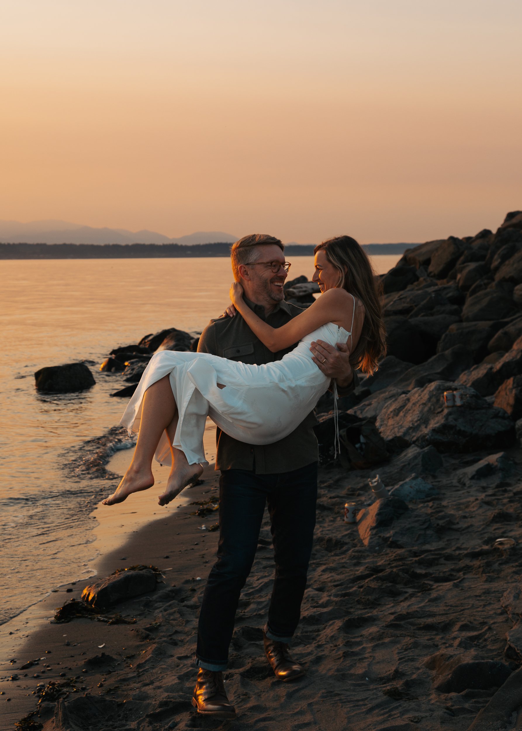 A man in dark pants and boots carrying a woman in a white dress on a beach at sunset. Discovery Park Beach Lighthouse elopement
