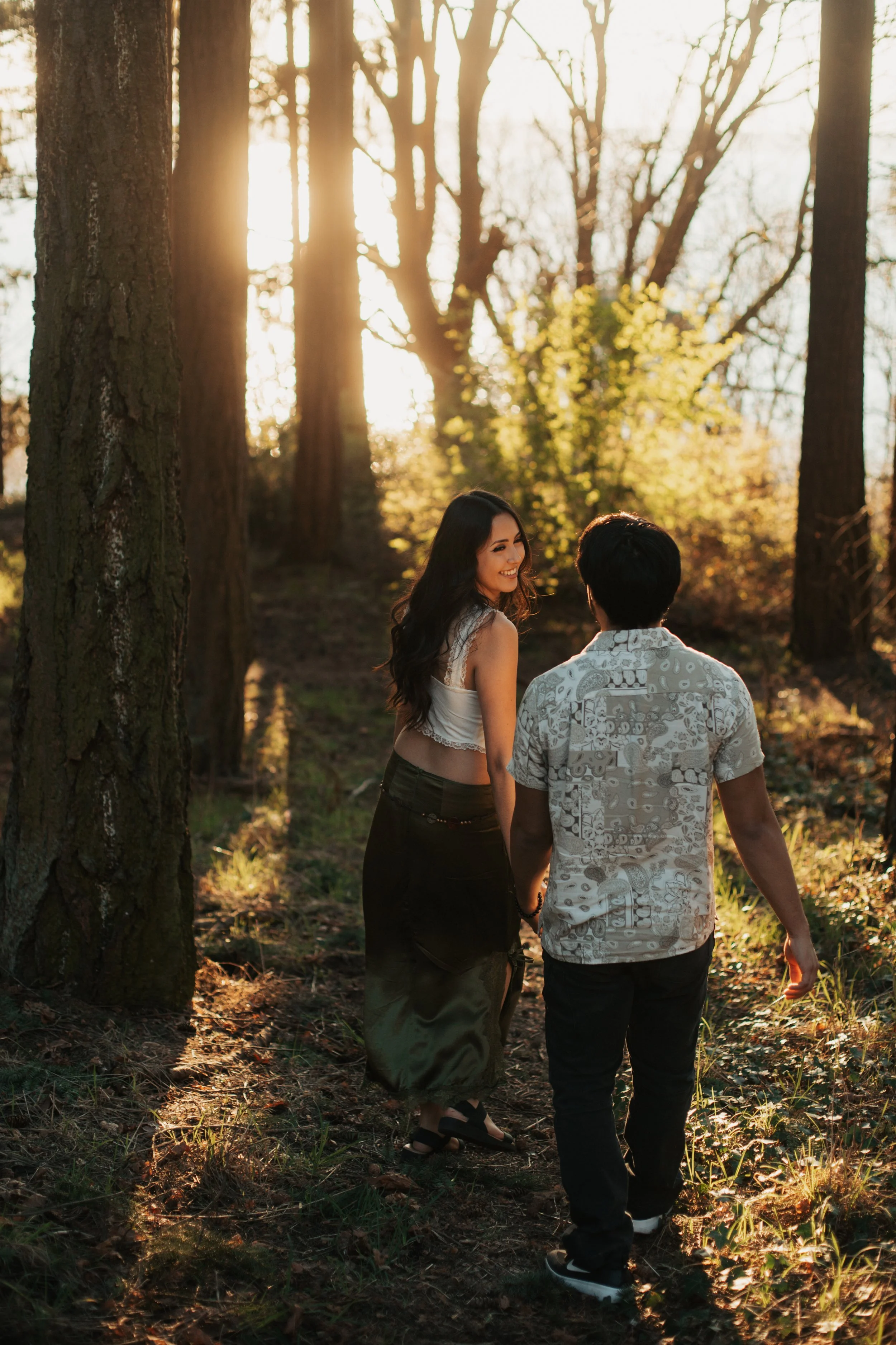 A young woman and man walking hand in hand through a forest at sunset, smiling and enjoying each other's company. Golden Gardens park engagement photos
