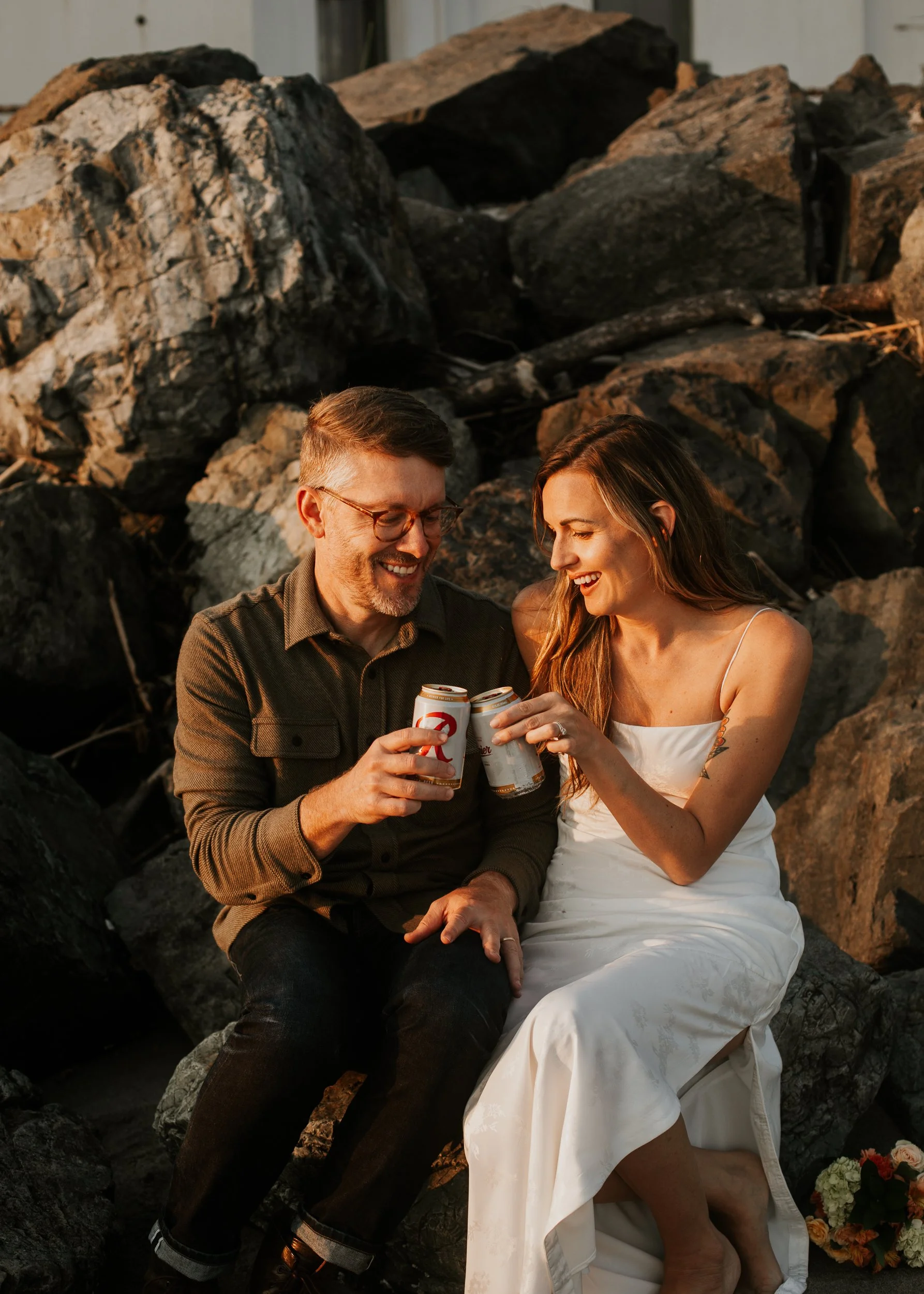 A couple sitting on rocks outdoors, sharing a drink and smiling at each other during sunset. Discovery Park Beach Lighthouse elopement