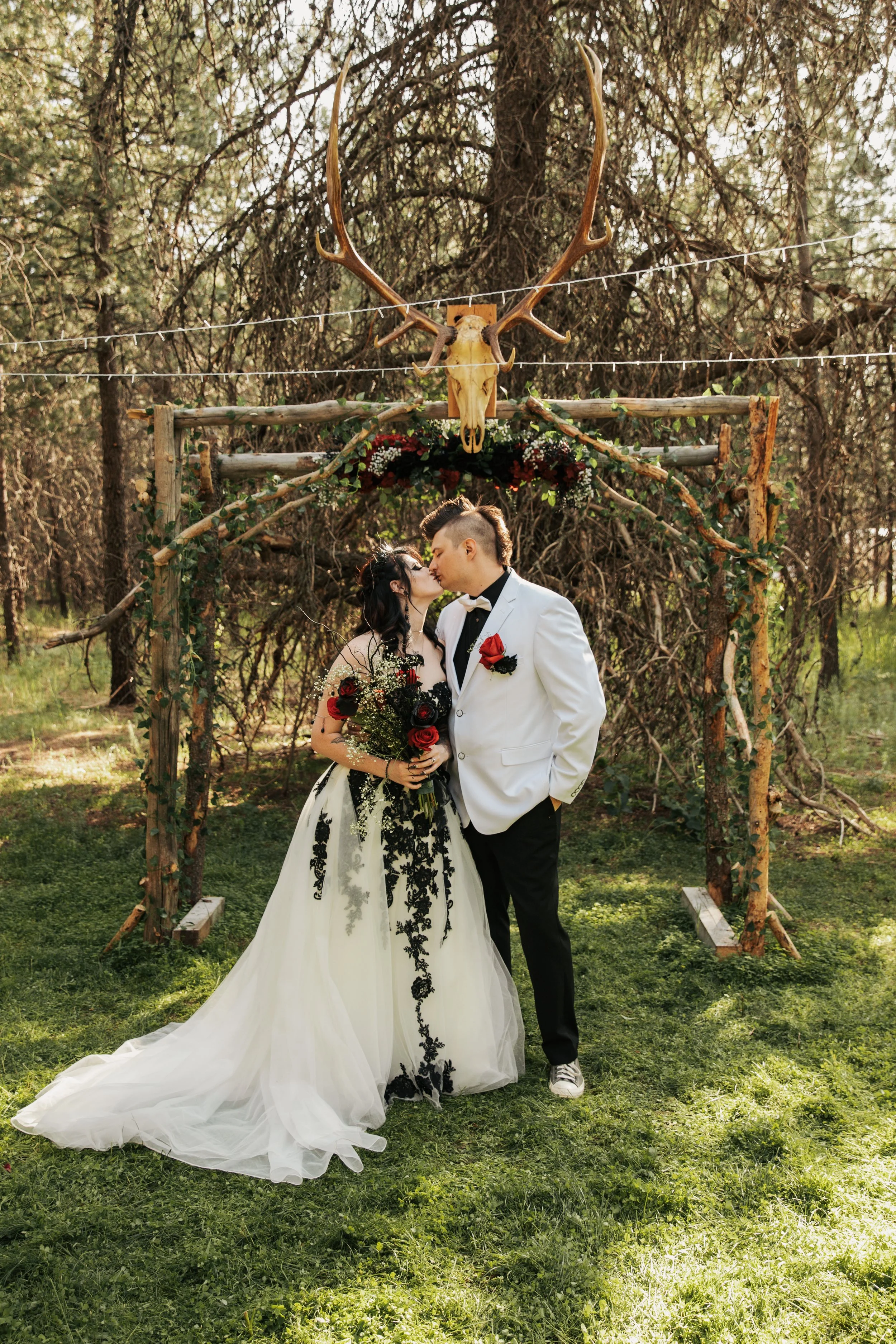 A bride and groom share a kiss at their outdoor wedding ceremony, with a rustic wooden arch decorated with flowers and a mounted deer skull with antlers above them. Athol Idaho wedding. Gothic wedding.