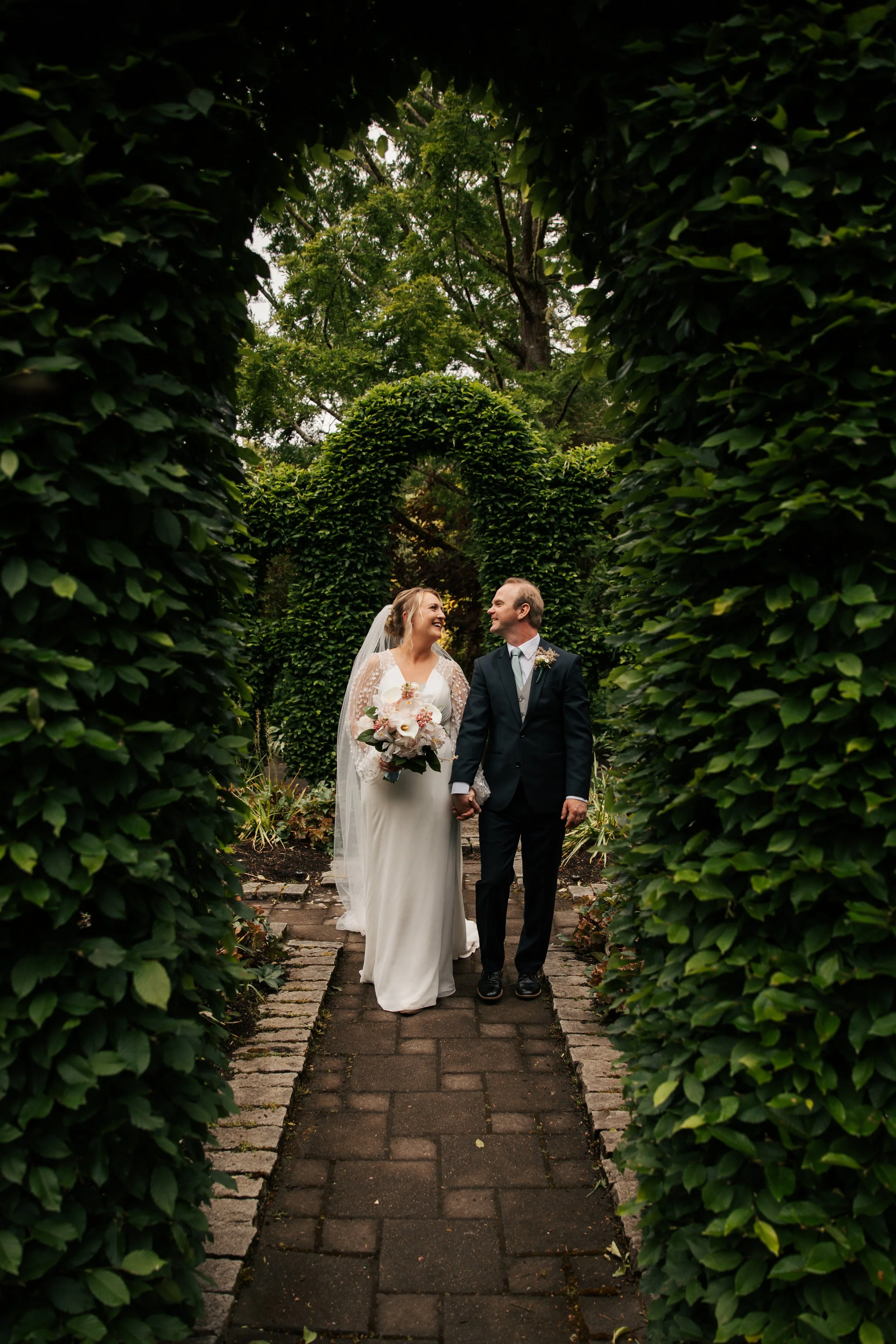 A bride and groom holding hands and smiling at each other while walking through a garden archway covered with lush green foliage. Heronswood Garden wedding Kingston Washington