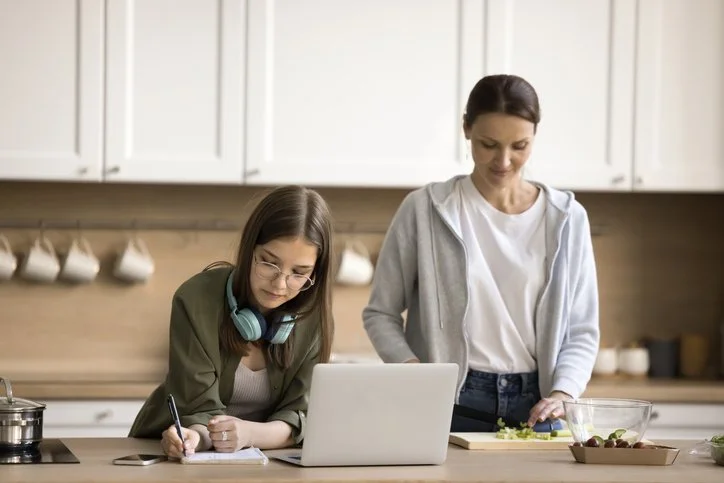 A mother chopping vegetables at a kitchen counter while her teenage daughter sits next to her doing homework.