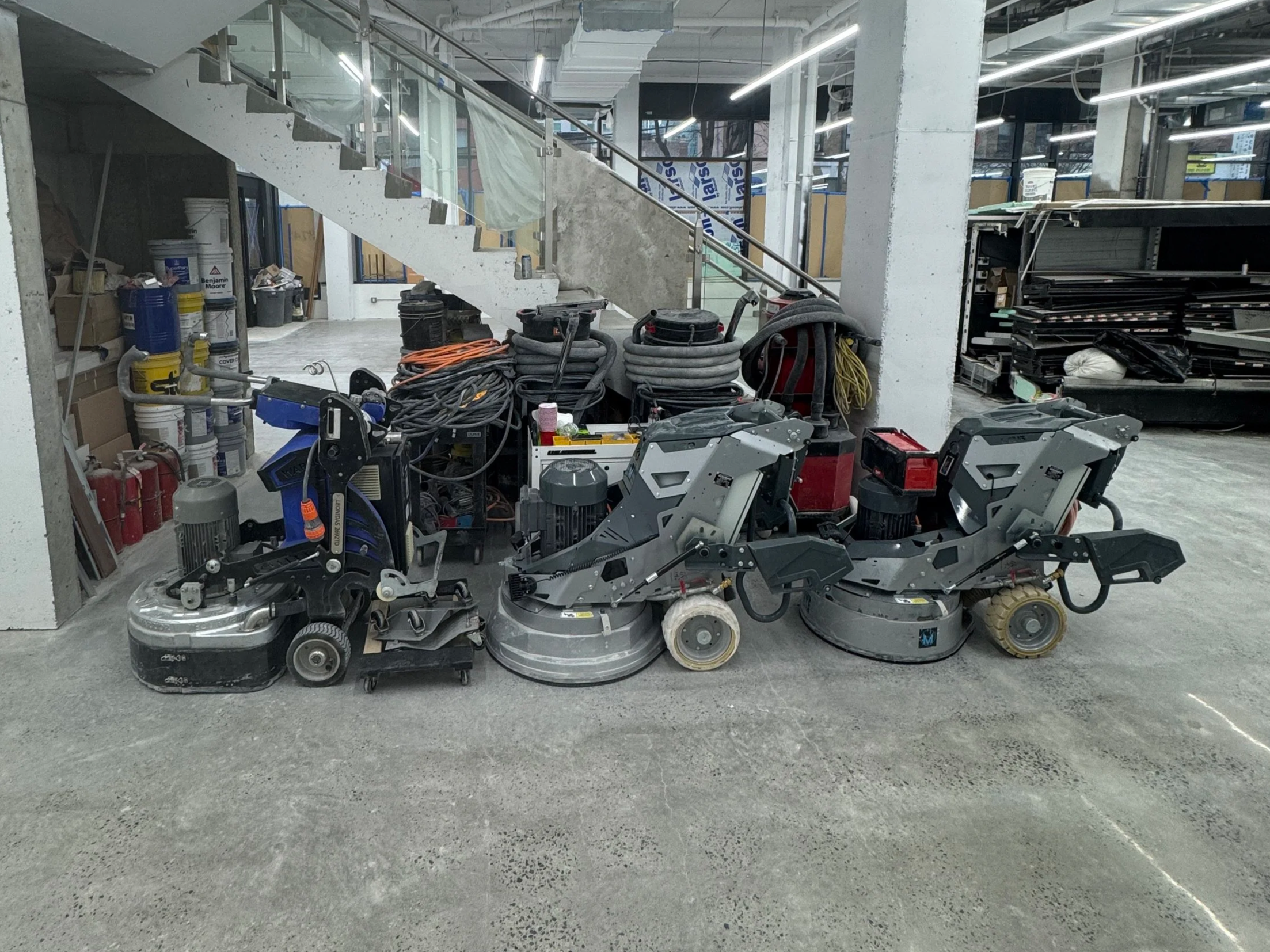 Construction site with equipment, tools, and materials for flooring or cement work, including riding floor saws and various stored supplies, in an unfinished building interior.