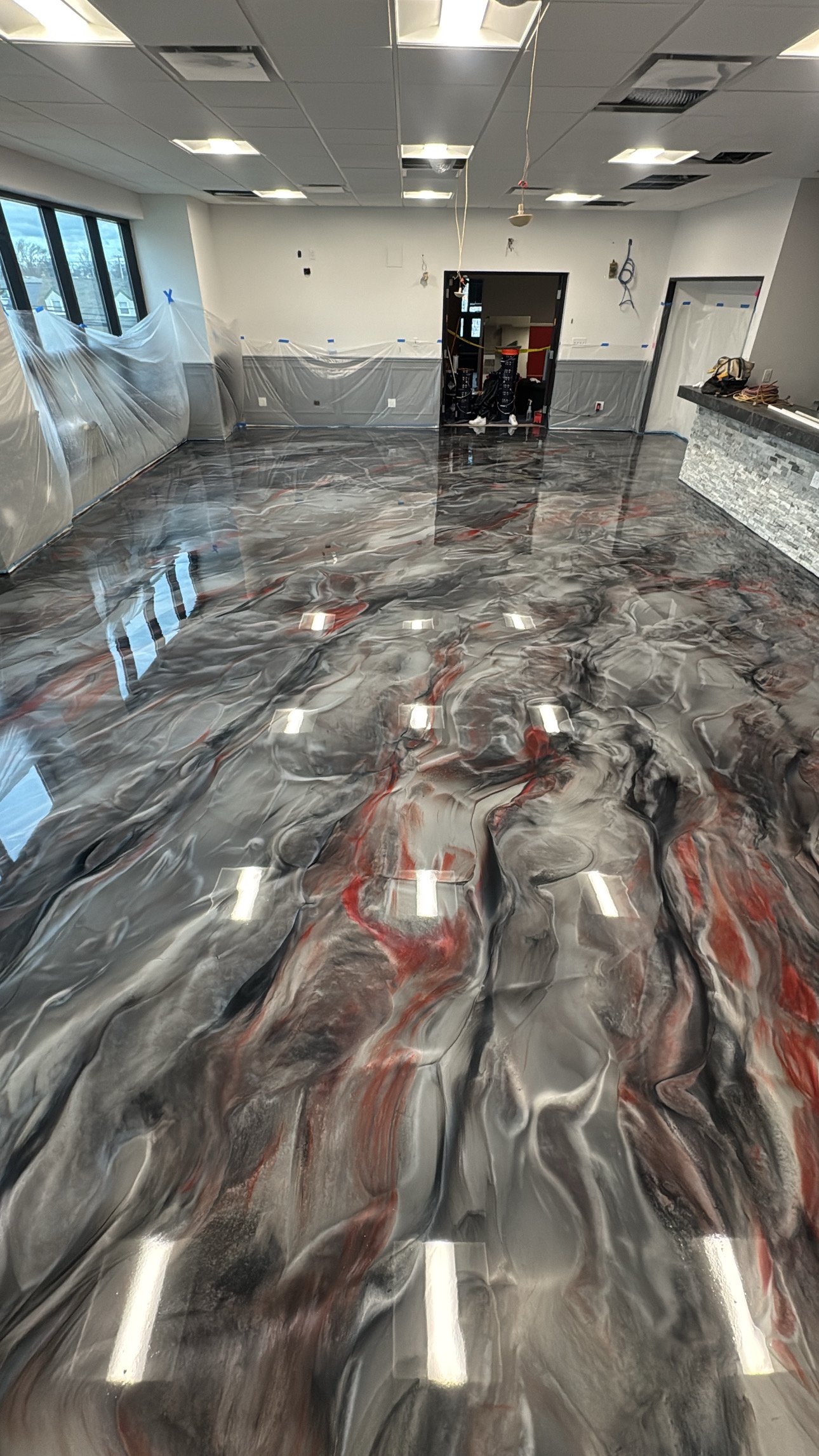 Interior of a room under renovation with a marbled epoxy floor, plastic sheeting on the windows, and unfinished ceiling with missing panels.