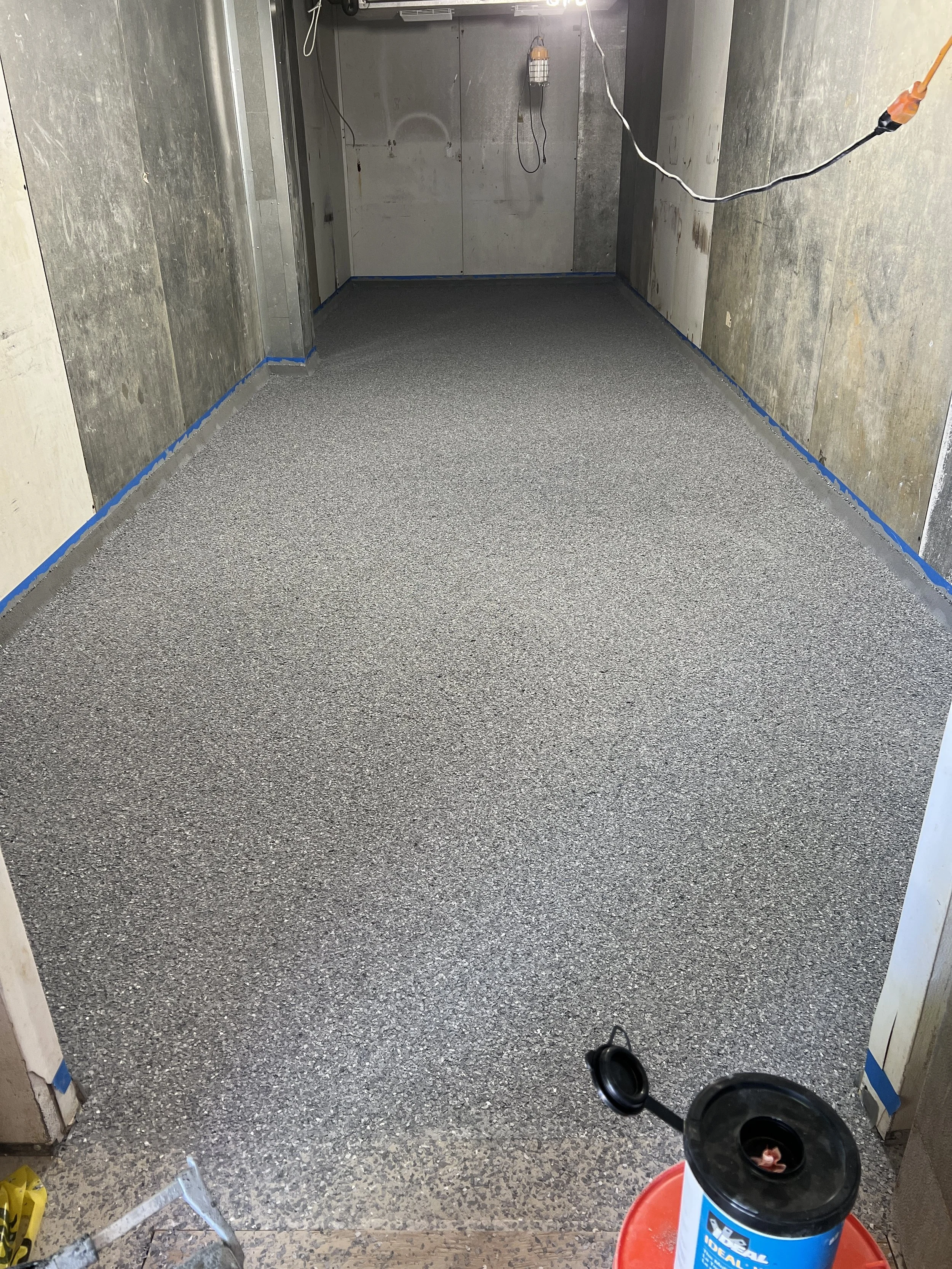 Room under construction or renovation with gray speckled flooring, unfinished drywall, hanging electrical wires, a propane heater, and construction supplies such as a yellow bag and a container of foam sealant.