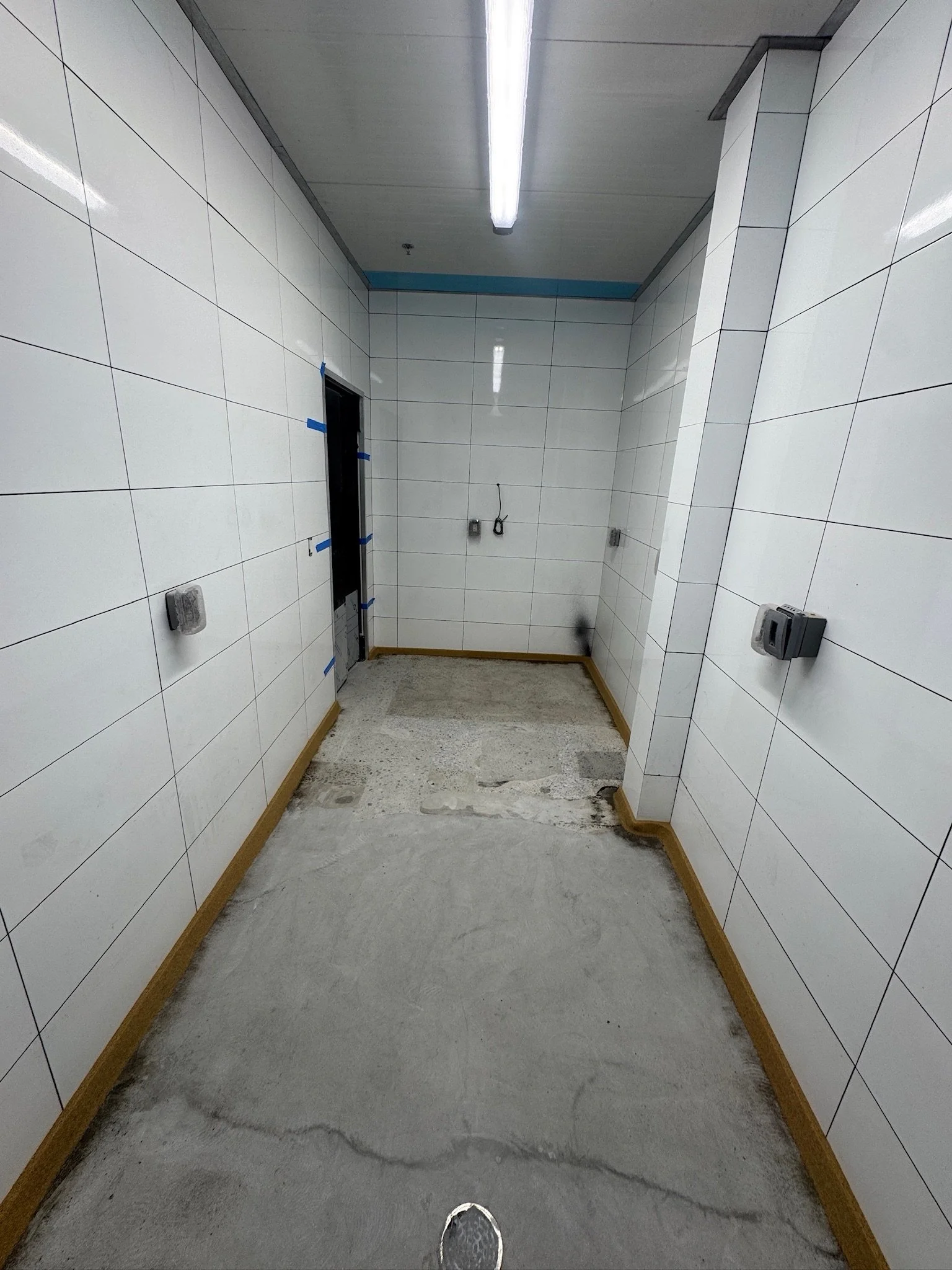An indoor hallway under construction with white tiled walls, a ceiling with fluorescent lighting, and a concrete floor with some wet patches. There are partially installed electrical outlets on the walls and an unsealed doorway.