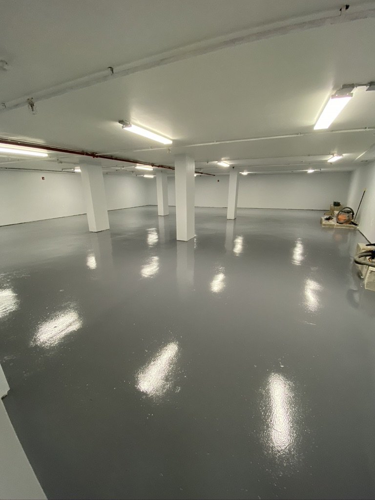 Empty underground parking garage with freshly painted gray floors and white walls, illuminated by fluorescent lights.