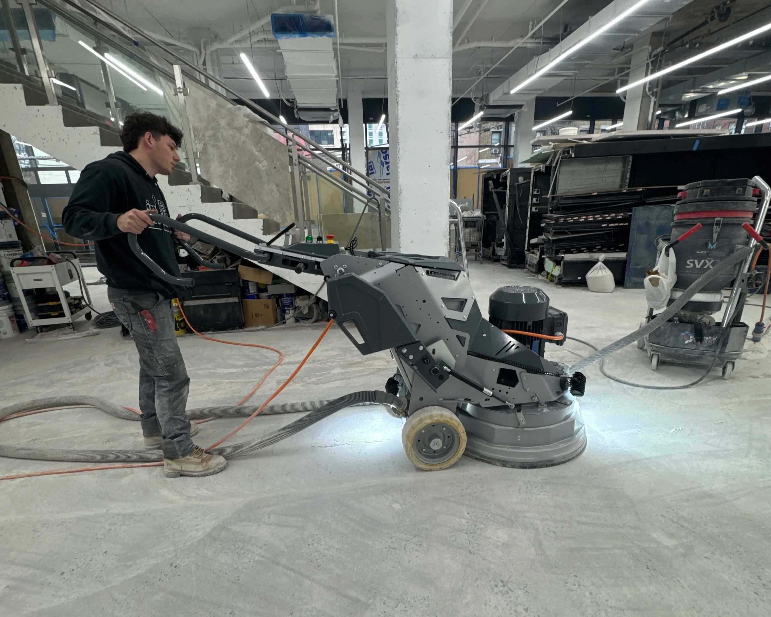 A person operating a large industrial floor grinder in a construction site or renovation area with concrete floors, machinery, and construction materials in the background.