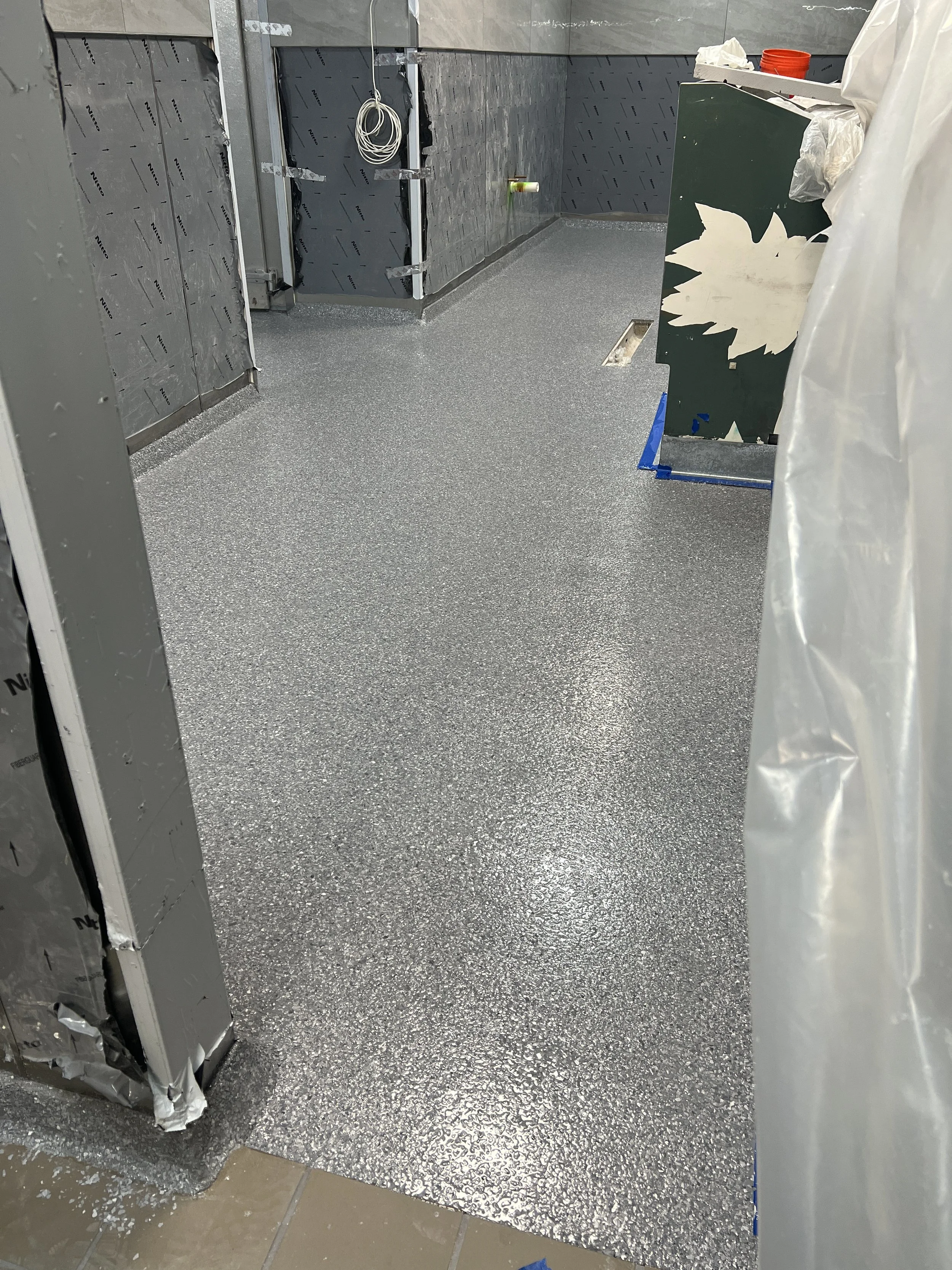 A room under construction with insulation, wiring, and unfinished walls. The floor has a speckled gray surface, and there are construction materials and tools present.