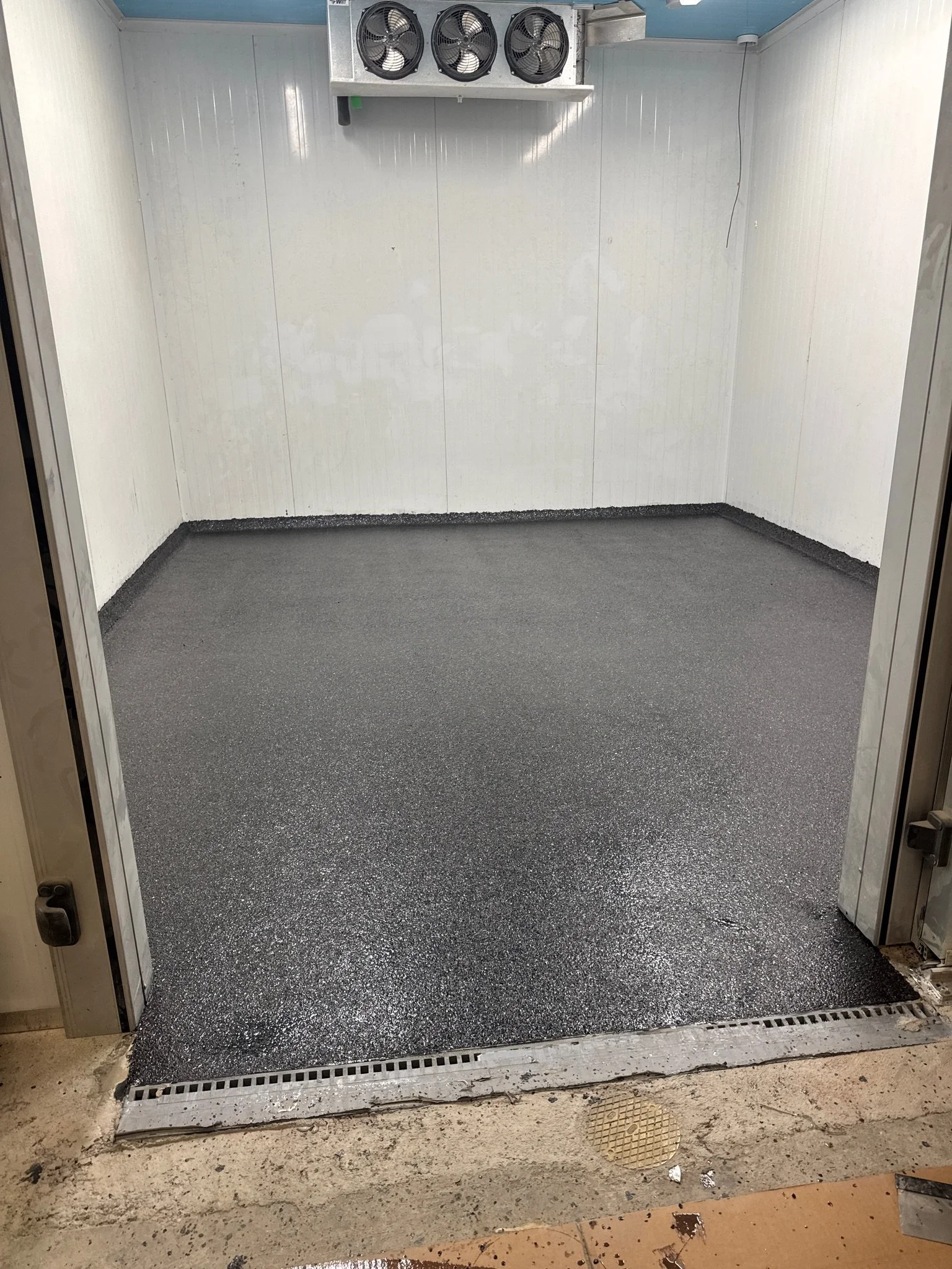 Empty elevator with black speckled flooring, white walls, and a fan ventilation system at the top. The elevator floor is shiny, and the corner of the elevator shows a small amount of dirt or debris on the ground outside.
