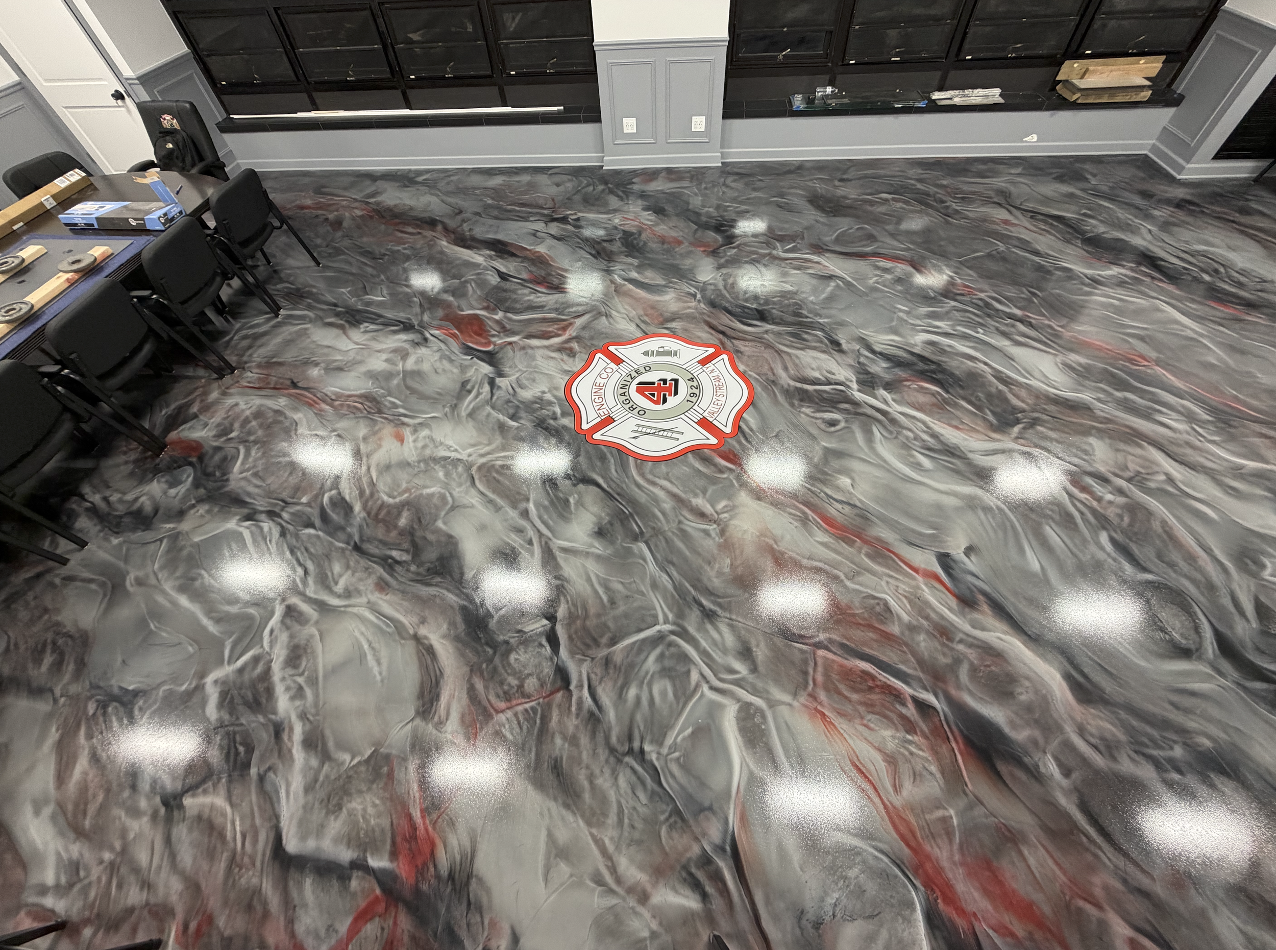 Industrial-style room with swirling red, black, gray, and white marble-patterned floor, black chairs, a black table with tools and materials, wall outlets, and a red and white fire department emblem on the floor.