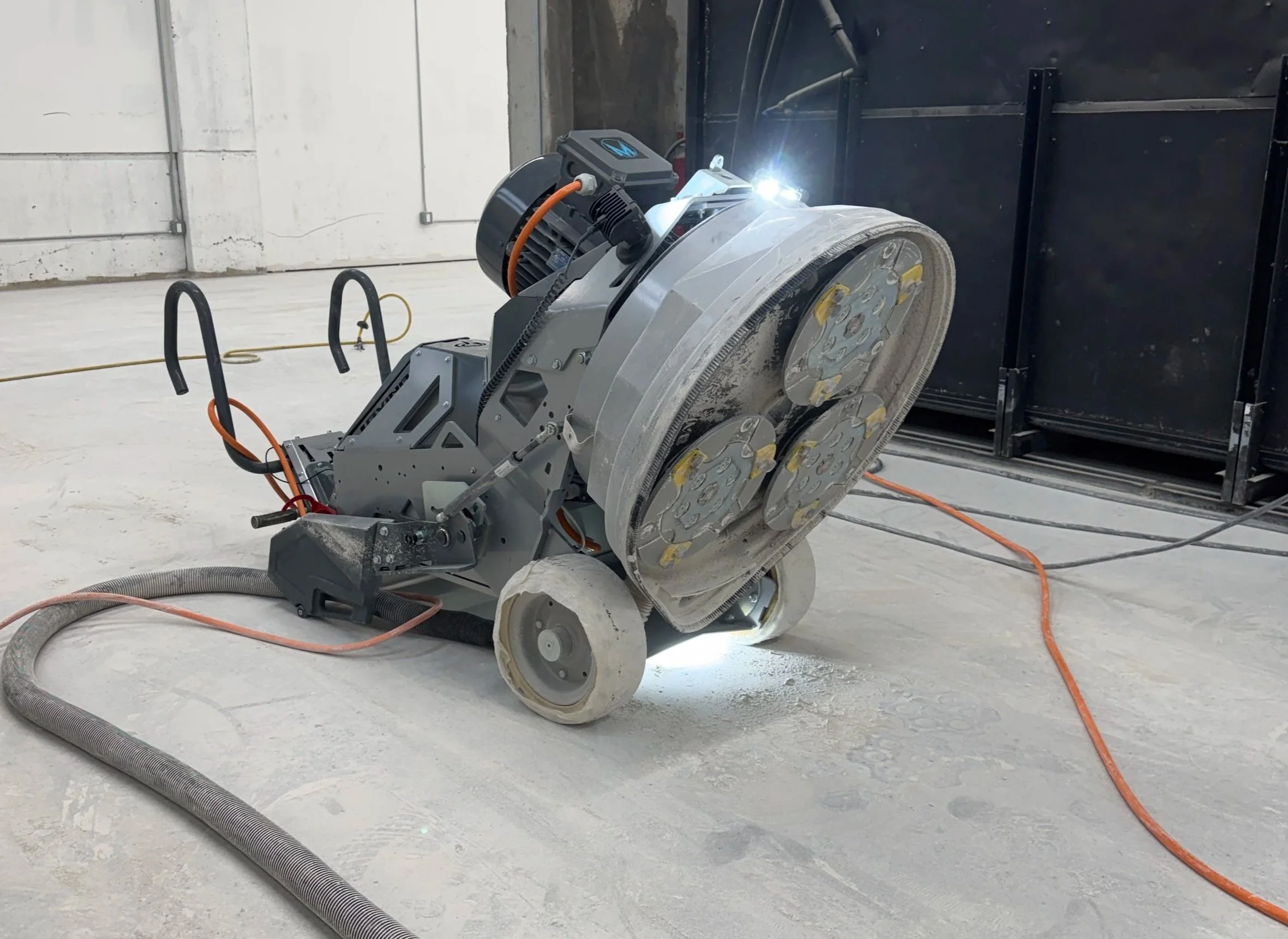 A large industrial floor saw cutting concrete in an indoor construction space, with dust and equipment cords around.