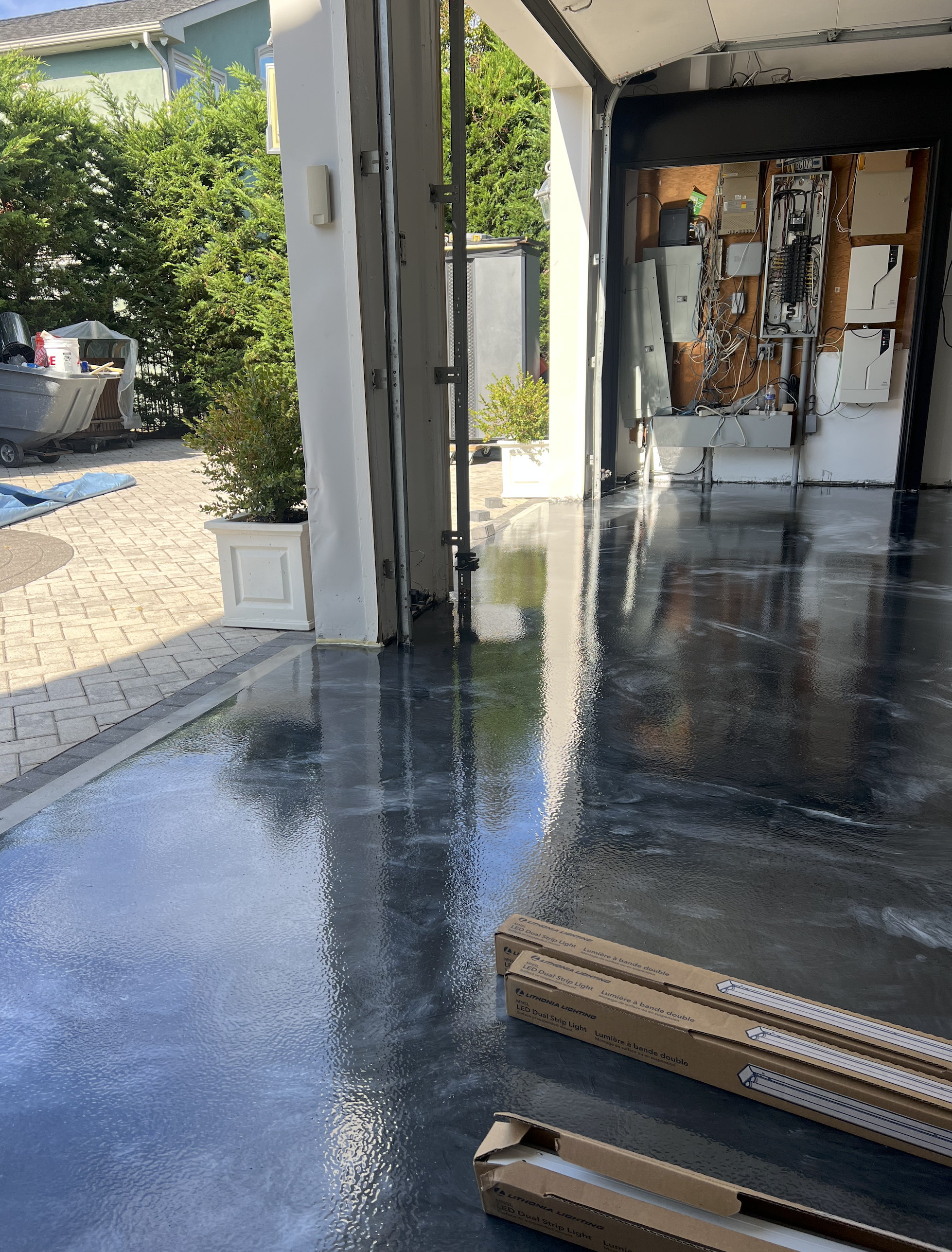 A garage with a partially opened garage door, revealing a shiny, wet polished concrete floor inside. Outside, there is a patio with pavers, a bush in a white planter, and a cart with gardening supplies. Inside the garage, on the floor near the entran