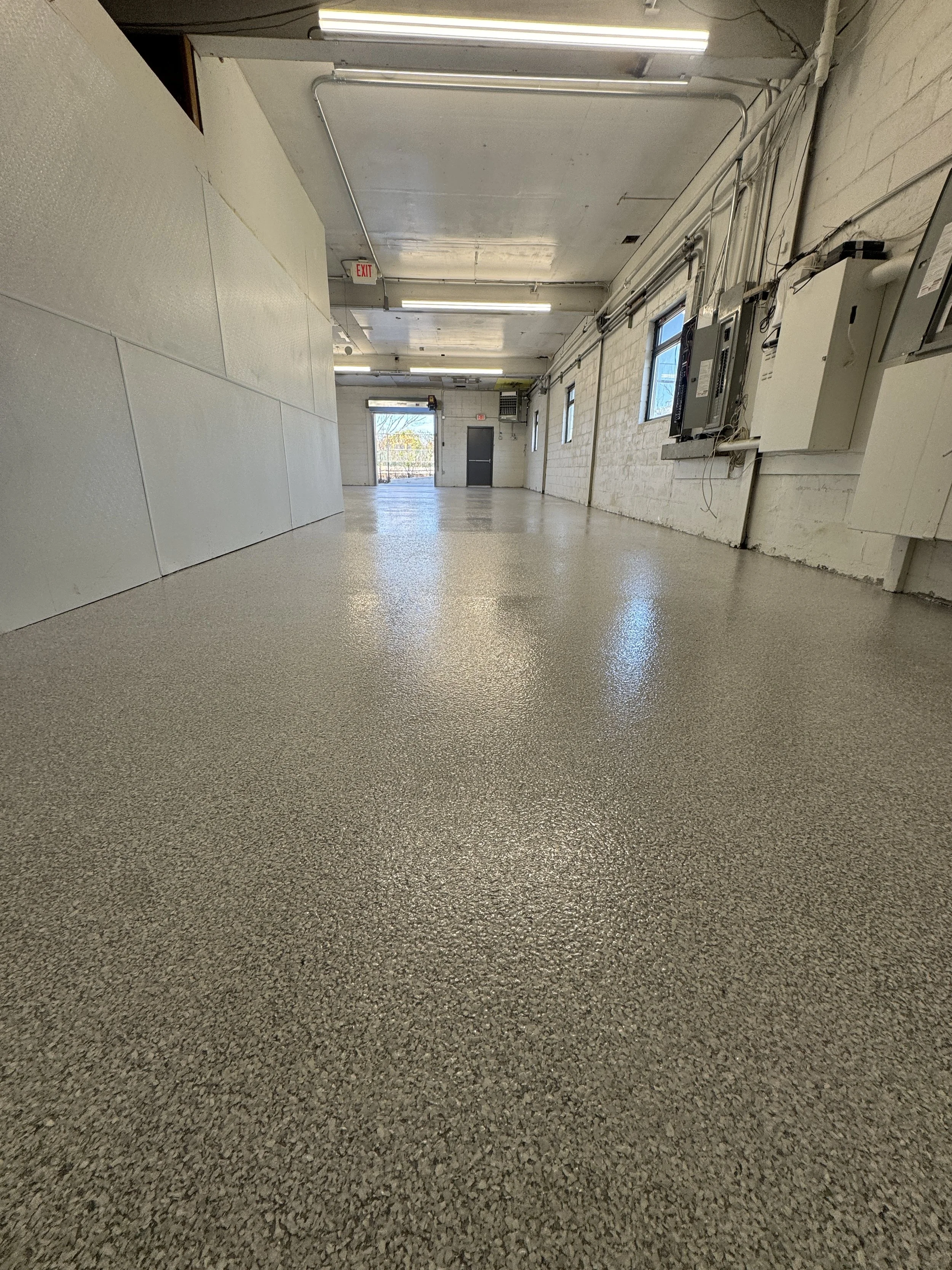 Empty commercial space or garage with textured gray flooring, white walls, and exposed wiring and electrical panels on the right wall. There are windows on the right wall and a glass door at the far end with an exit sign above it.