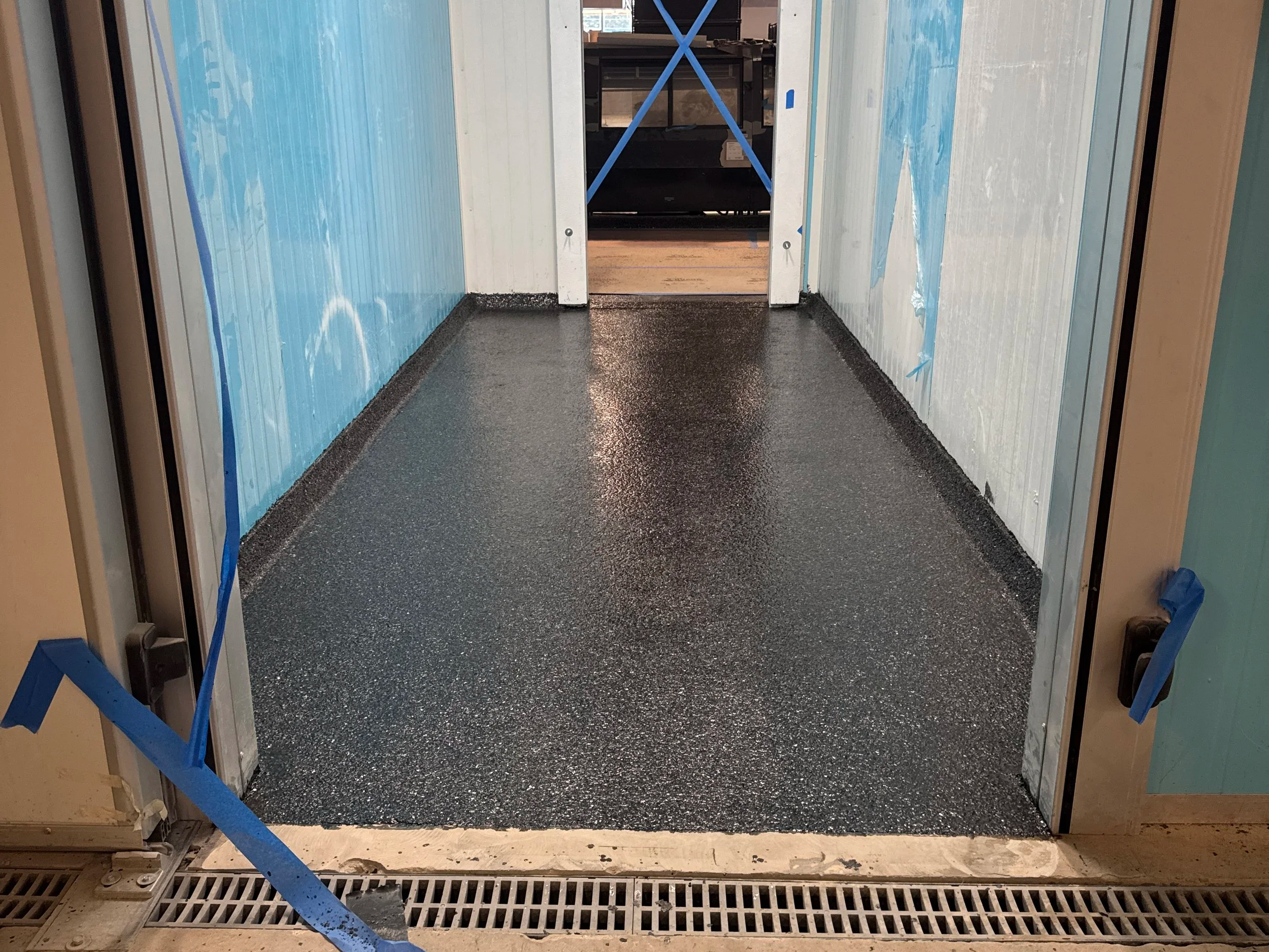 Freshly coated black epoxy floor in a hallway with white walls and a door opening at the end, taped off with blue painter's tape.