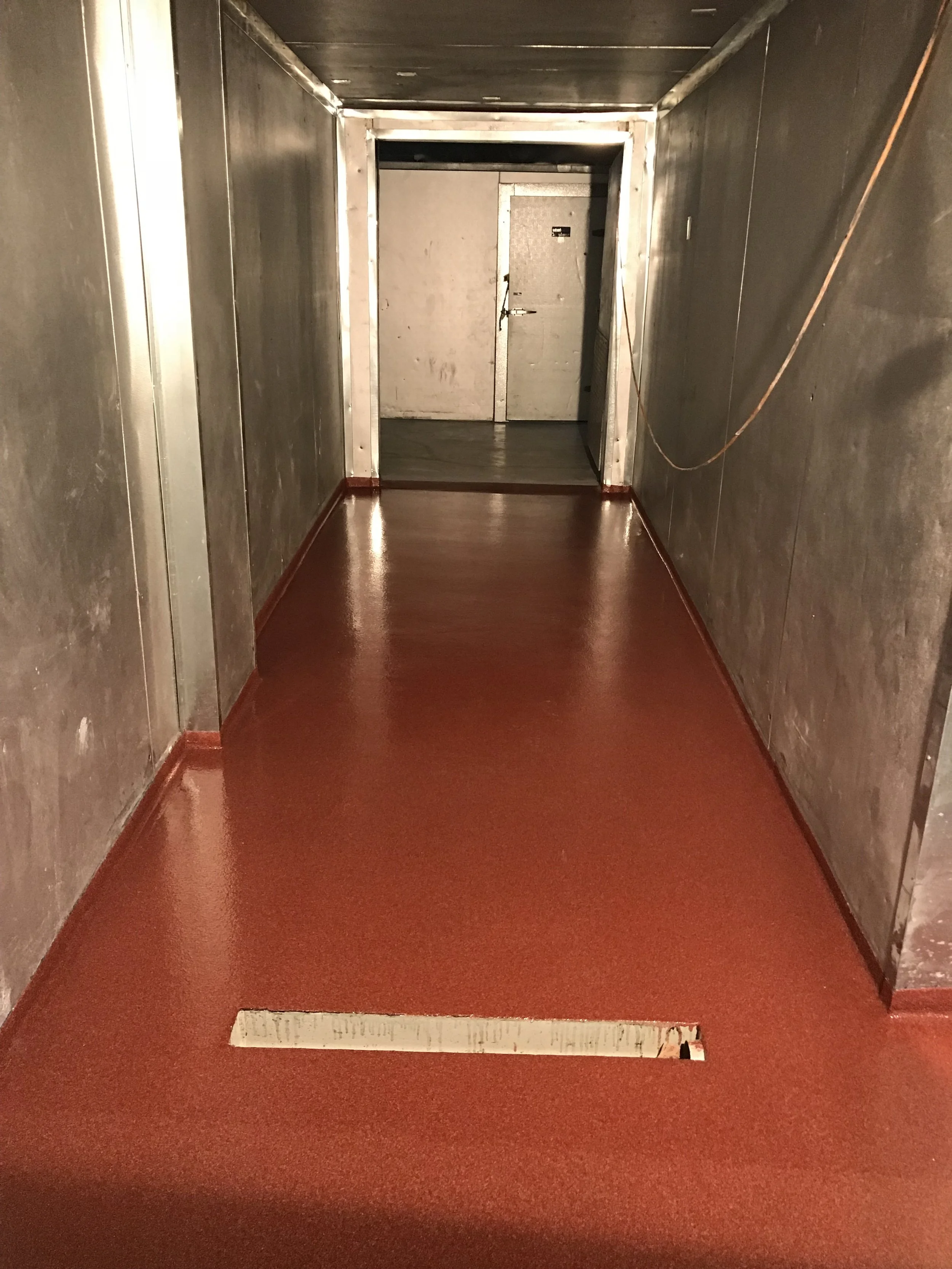 Empty hallway with red flooring, gray walls, and a closed metal door at the end with a lock and a small window.