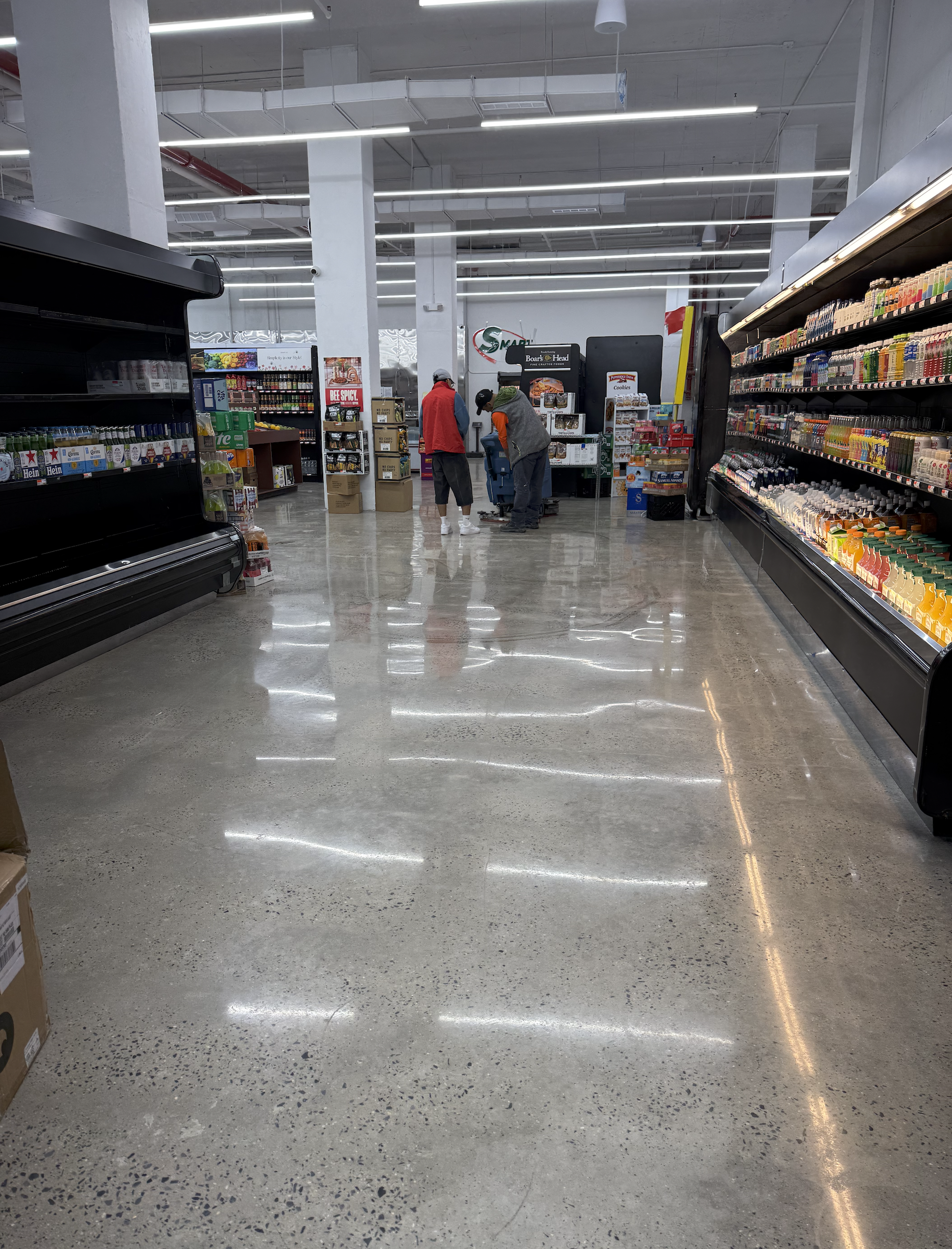 View of a grocery store aisle with shelves on either side stocked with various products, two people shopping near the checkout area, concrete flooring, and bright fluorescent lighting.