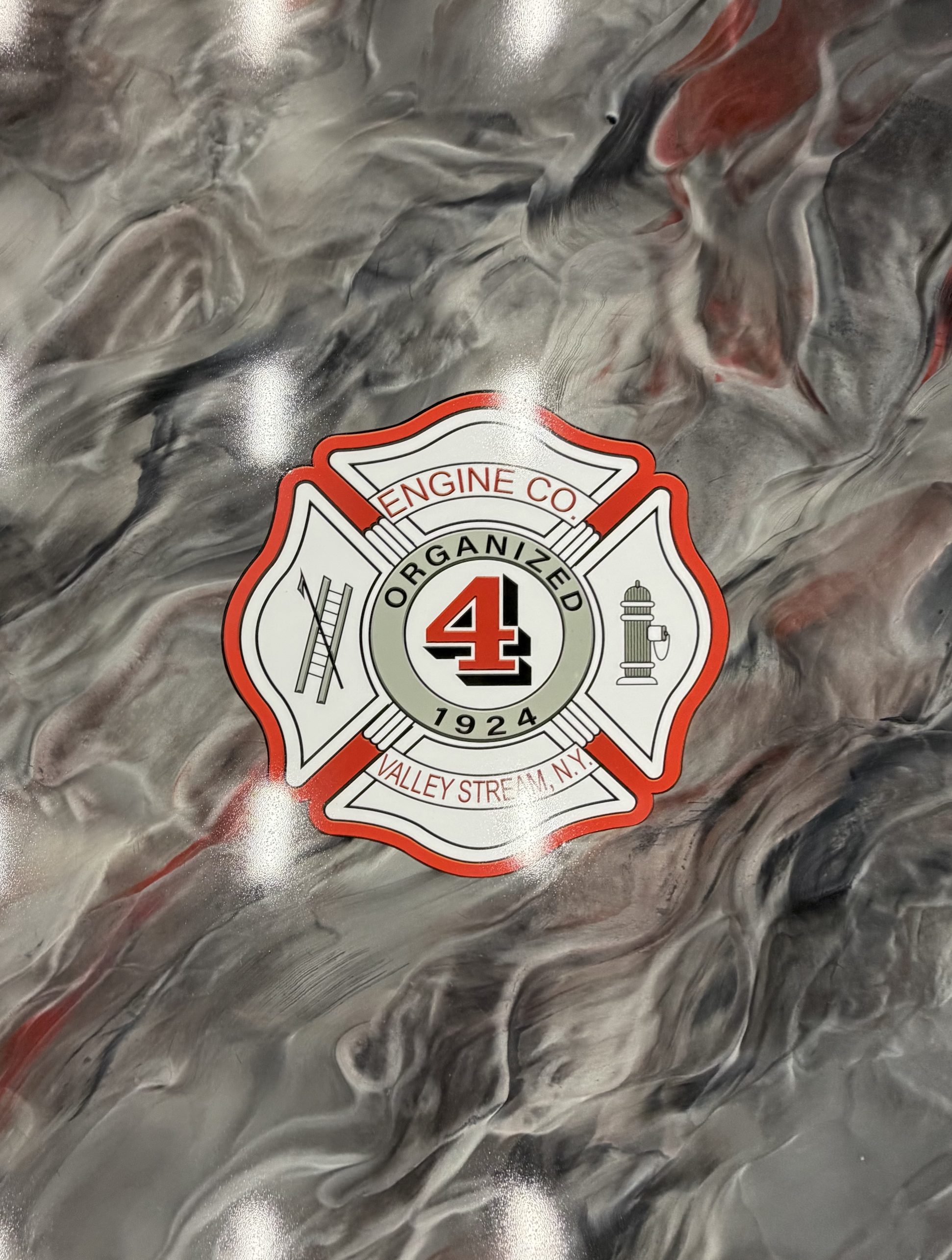 A metallic surface with a firefighter emblem sticker in the center, featuring a red border, a badge shape, and symbols of a ladder, fire hydrant, and a sprinkler, with a marbled gray and red background.