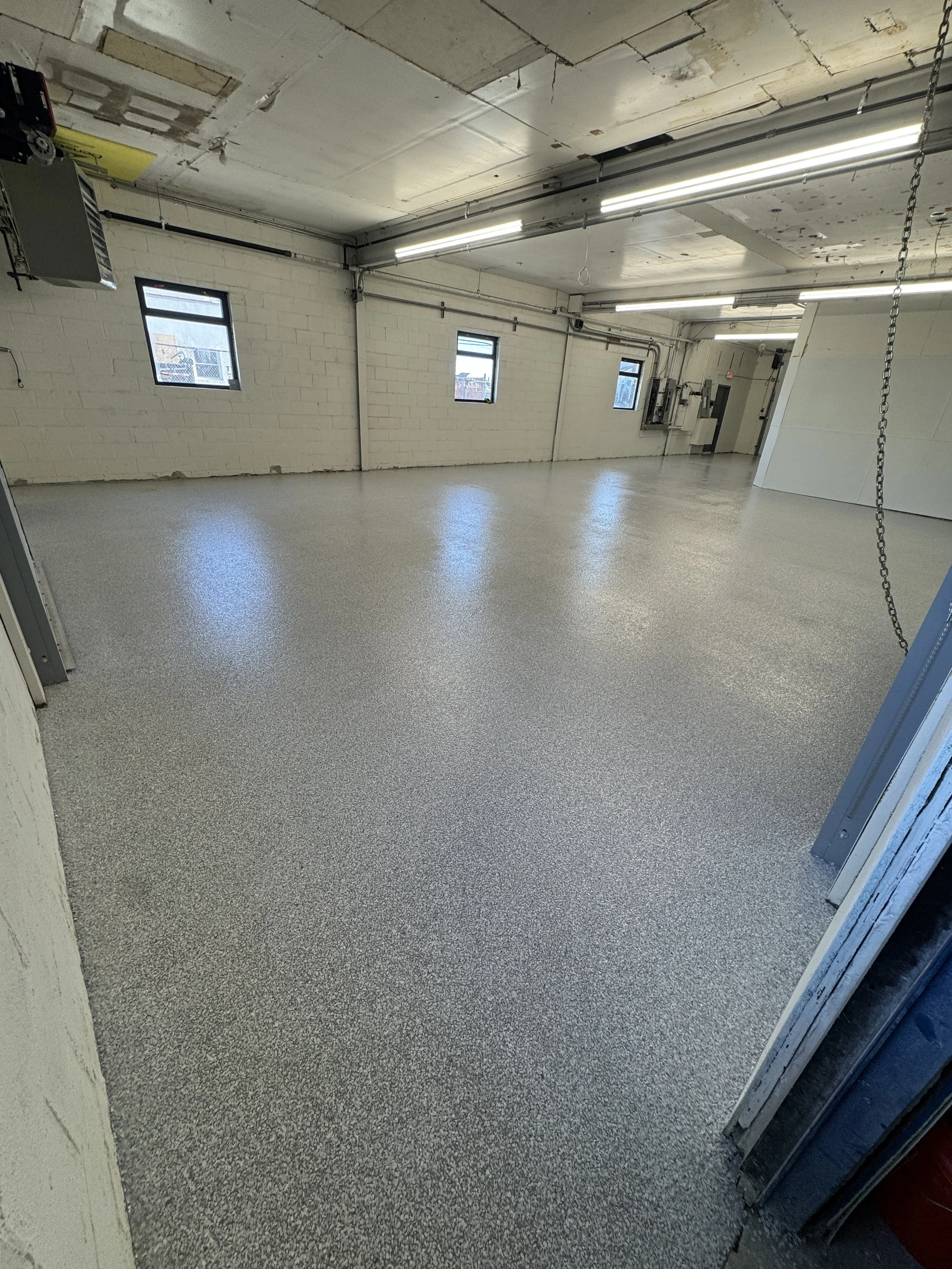 Empty industrial garage or workshop space with white brick walls, small windows, fluorescent lighting, and a speckled gray epoxy floor. There is some exposed piping and an electrical panel on the wall.