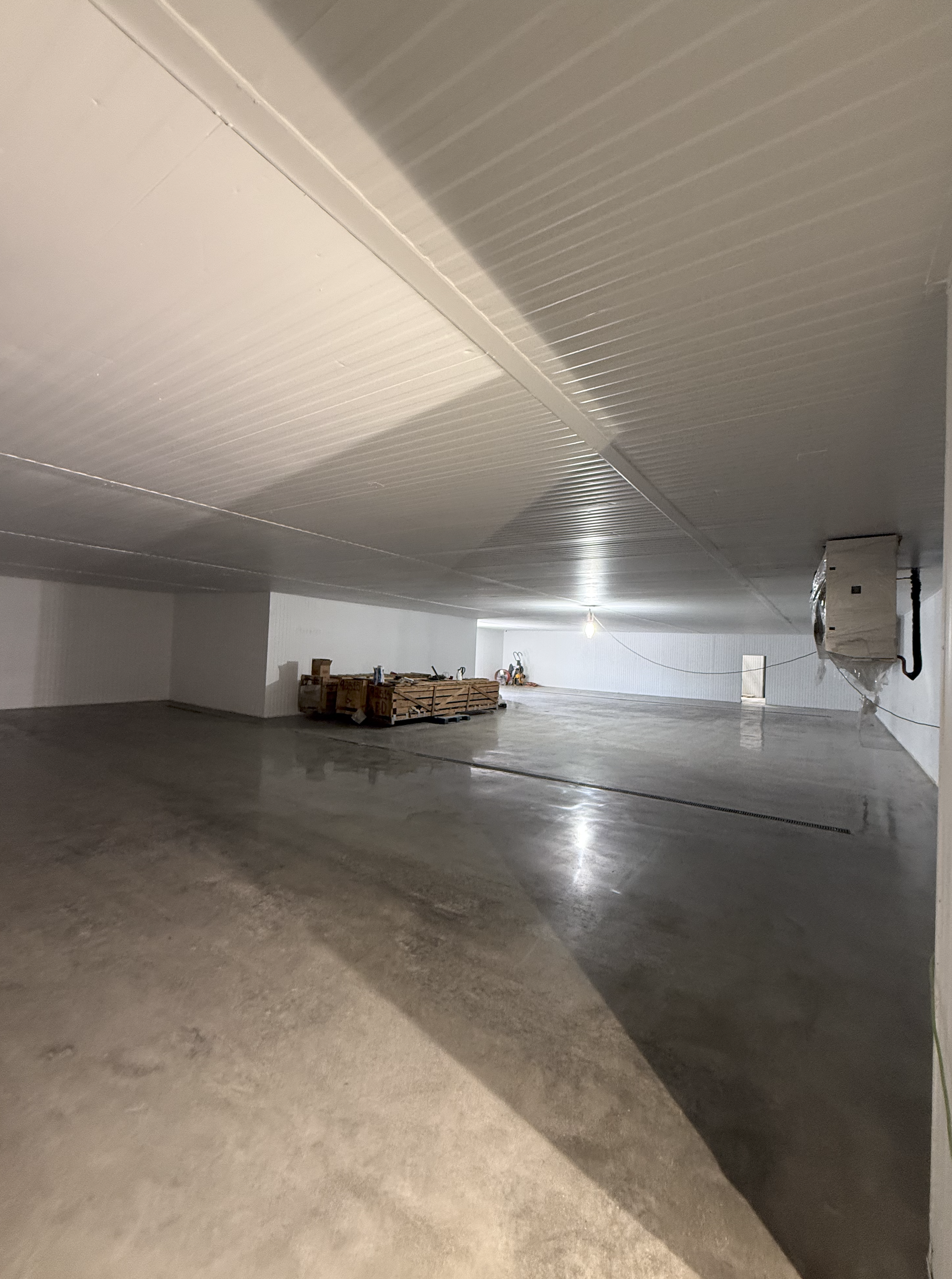 A large, open indoor space with a bushed concrete floor, white walls, and a ceiling with visible beams. There are some construction materials and tools near the back wall, and a single light bulb hanging from the ceiling.